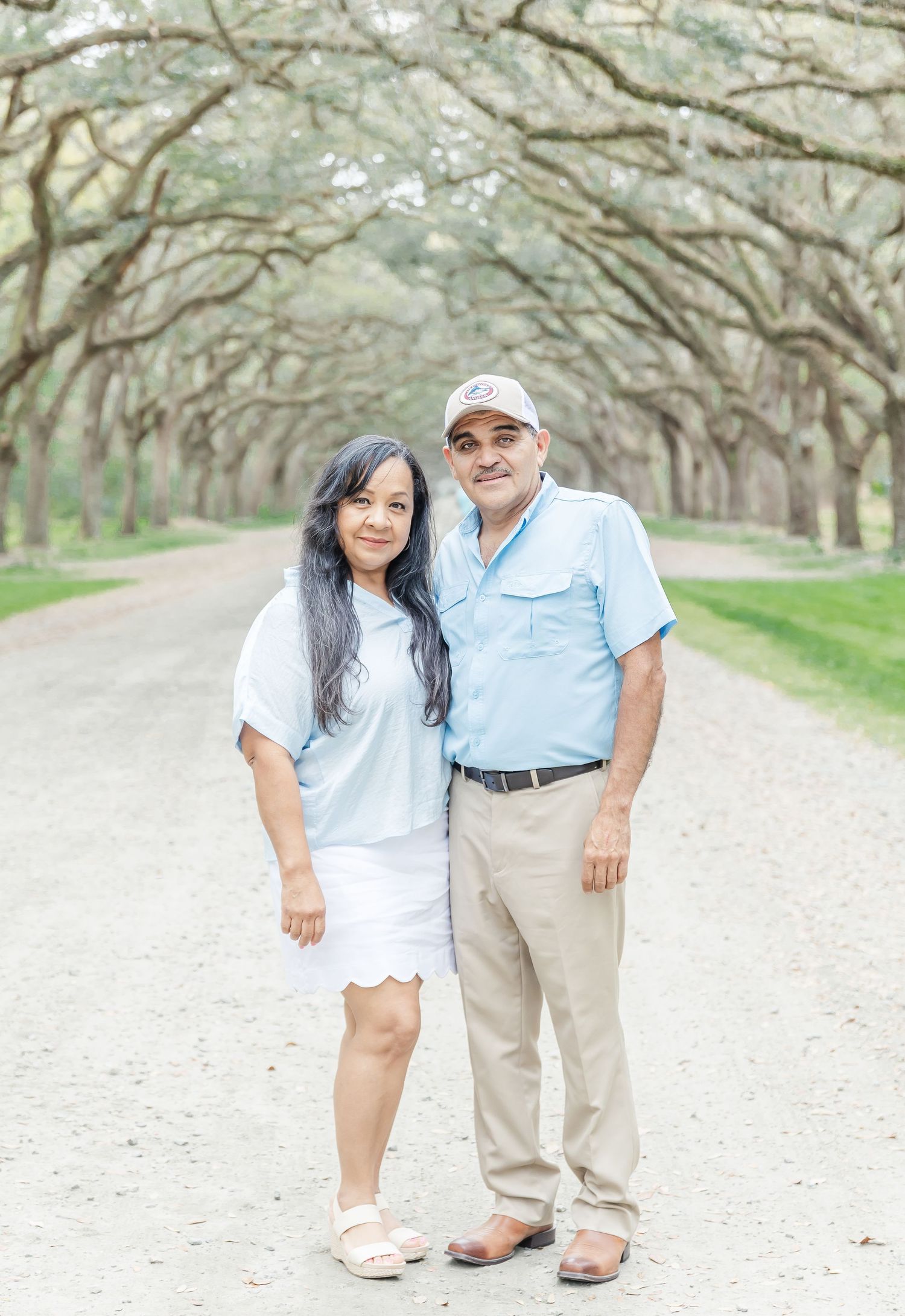 Couple standing together on dirt path under canopy of oak trees in casual summer attire.
