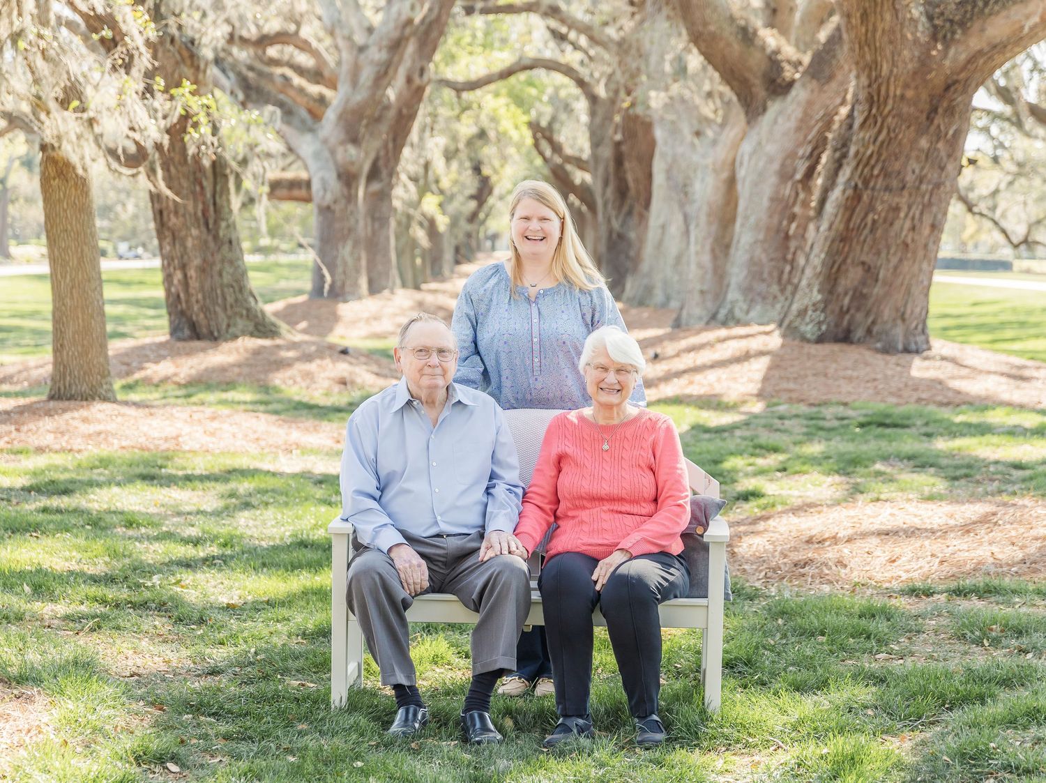 Three people posed on white bench under canopy of Spanish moss covered oak trees.
