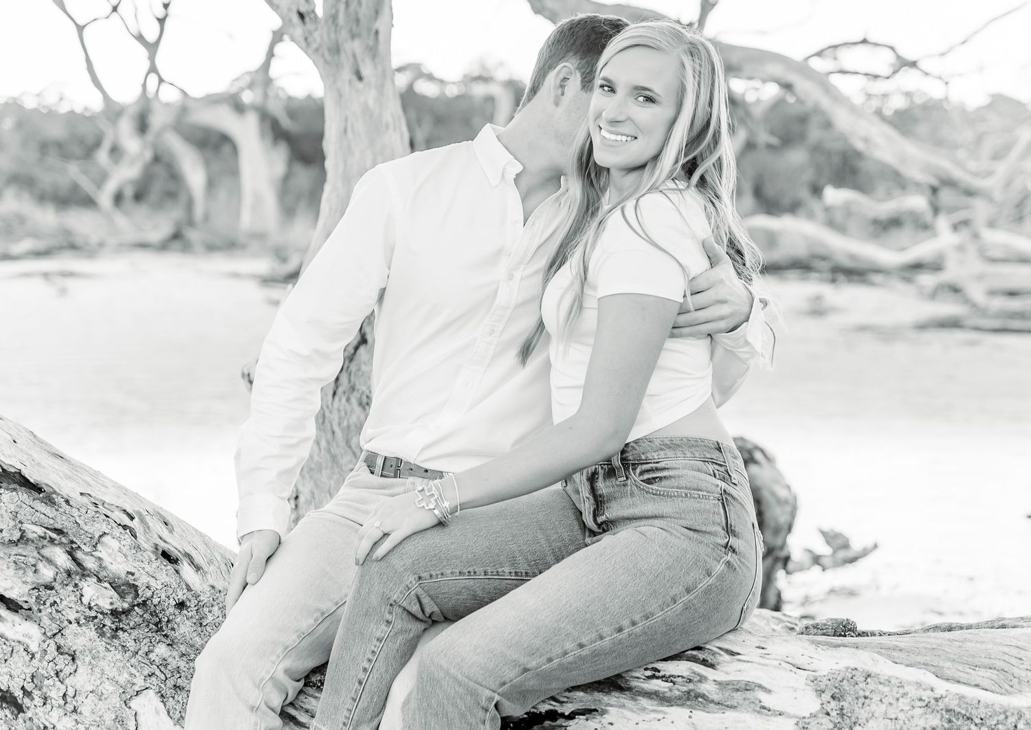 Black and white photo of a couple sitting together on a fallen tree trunk by the water.