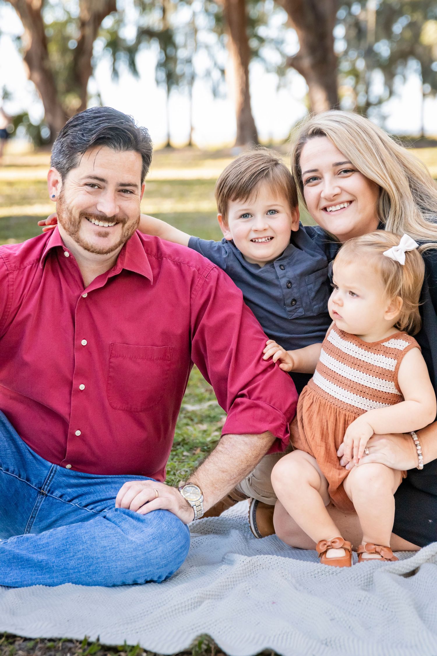 Happy family of four sitting together on blanket under oak trees.