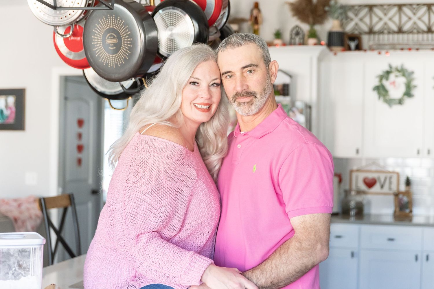 A couple in matching pink outfits poses together in their kitchen with hanging pots above.