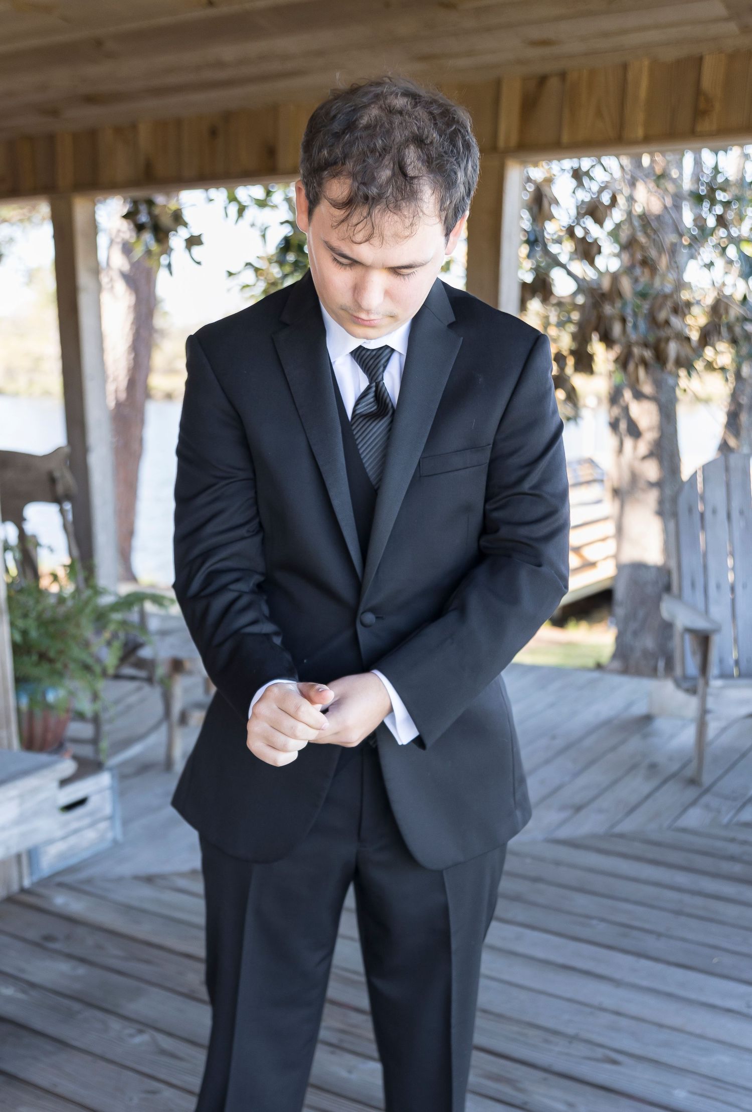 Groom in black suit adjusts buttons while standing on wooden porch.