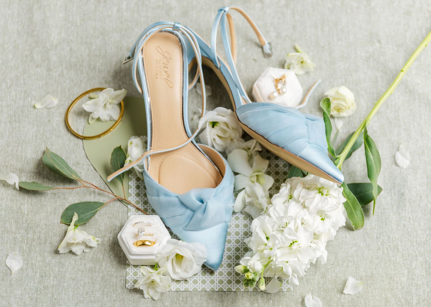Light blue slingback heels with white floral arrangements and delicate eucalyptus leaves on a neutral gray background.