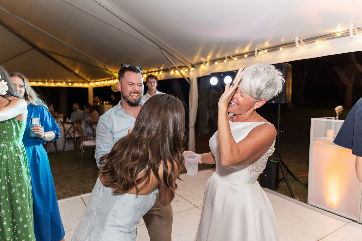Guests mingle and dance under a tent at an outdoor evening celebration with string lights overhead.