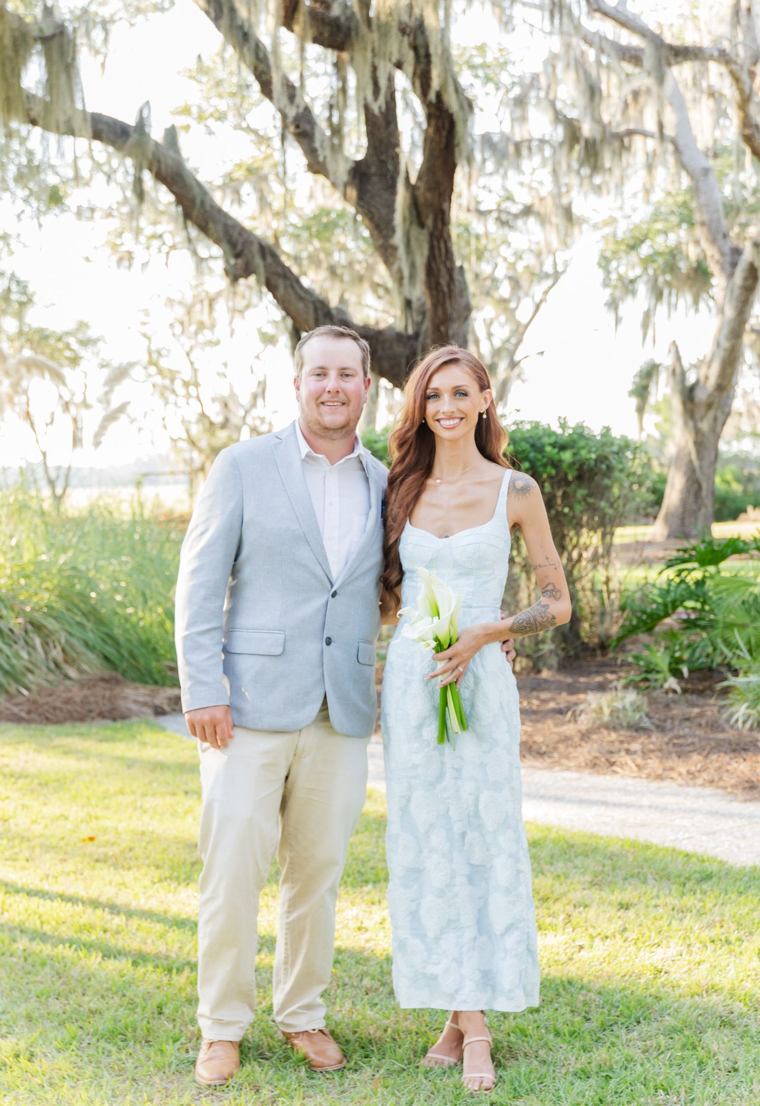 A couple poses together outdoors under moss-draped oak trees on a sunny afternoon.