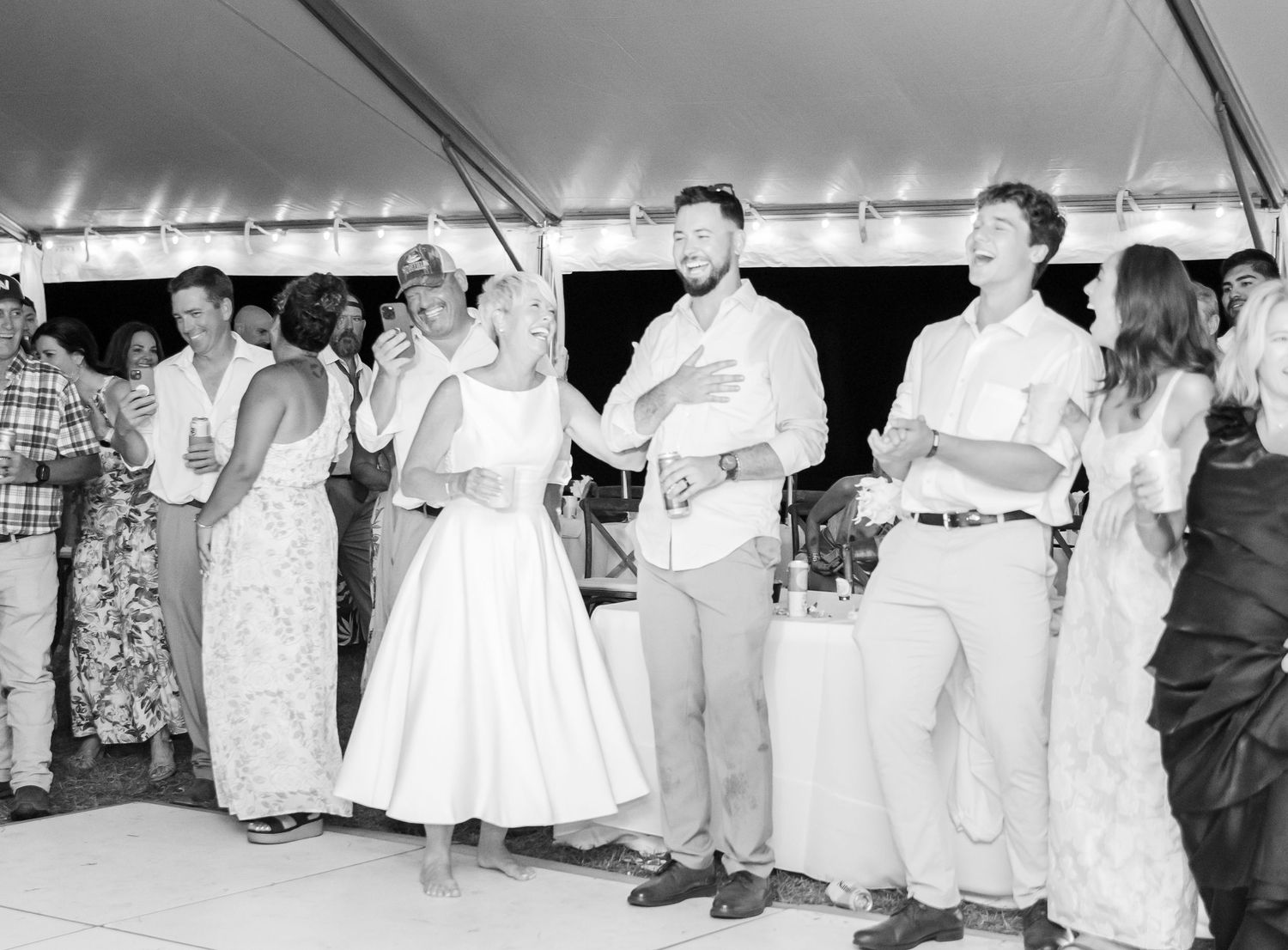A black and white panoramic photograph shows a group of people seated outdoors under a tent during an event.