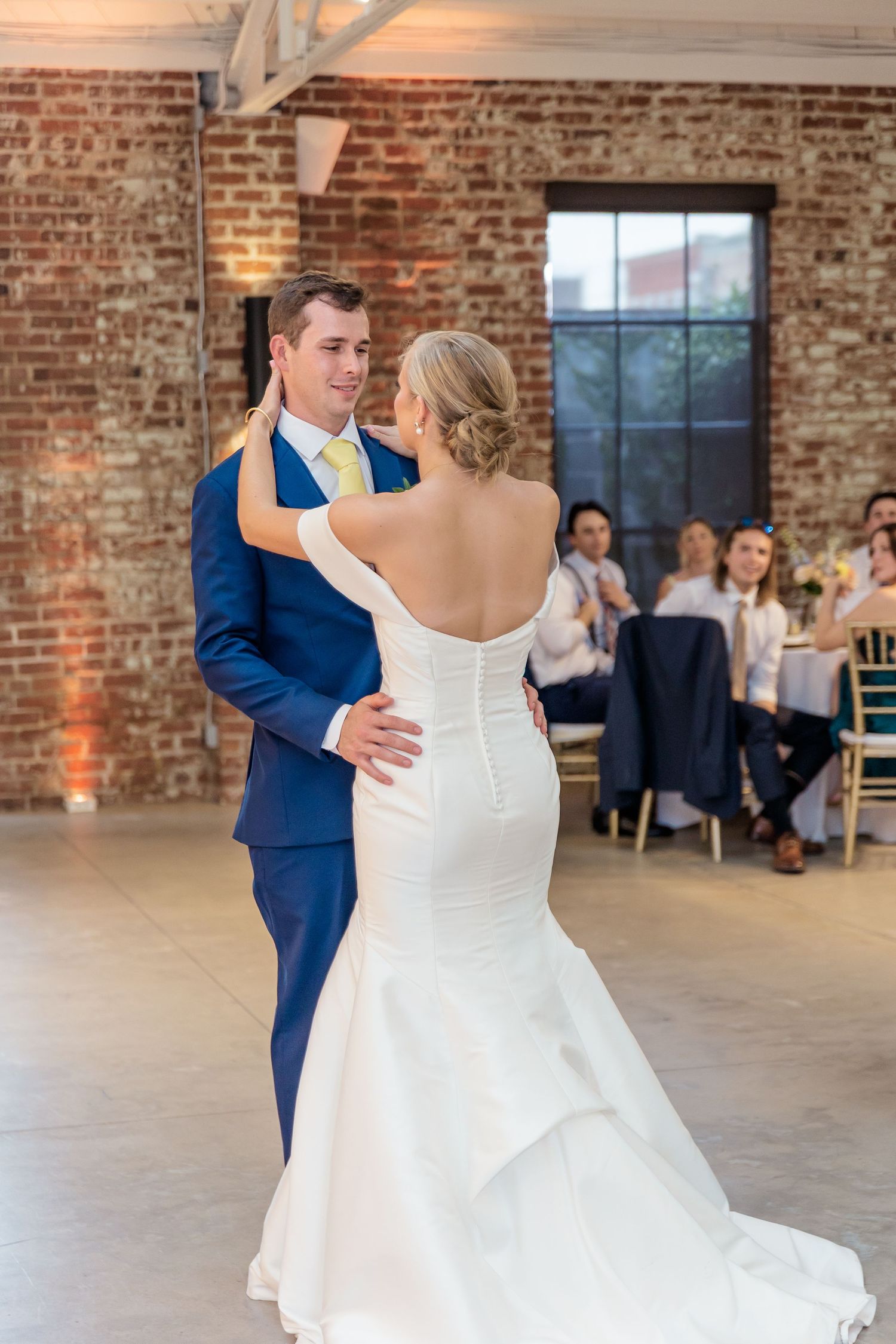 A couple shares their first dance in a rustic brick venue with guests seated at tables in the background.