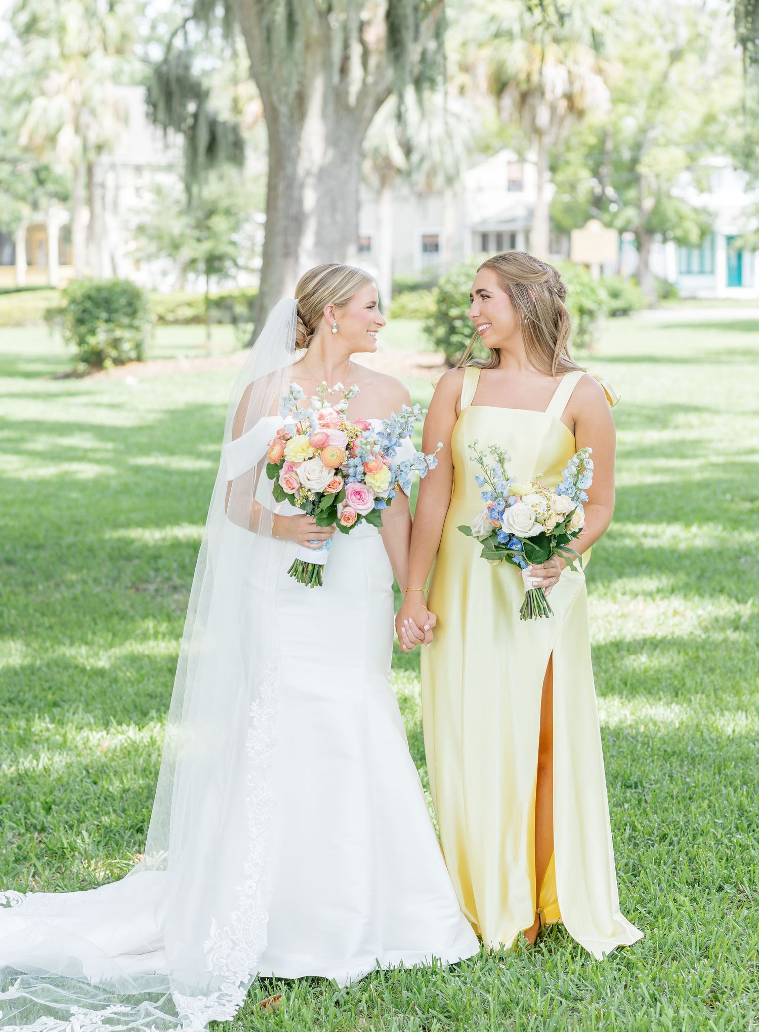 Bridesmaids in pale yellow dresses and bride in white gown walk together on sunlit garden lawn.