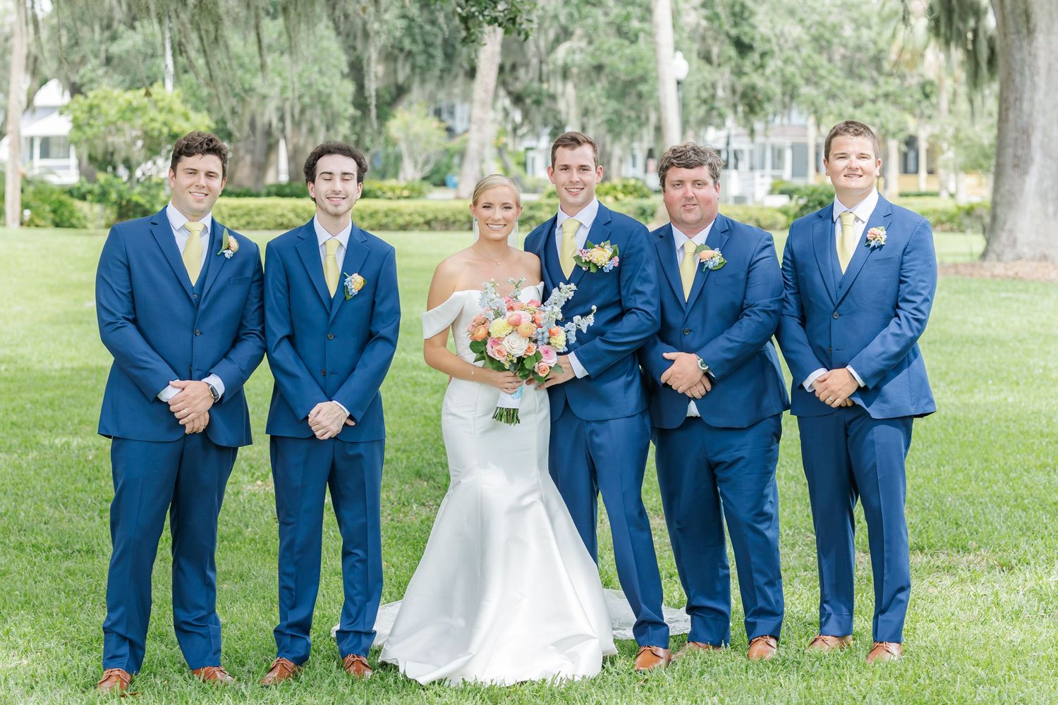 Wedding party in bright blue suits and white dress pose together on green lawn during outdoor celebration.