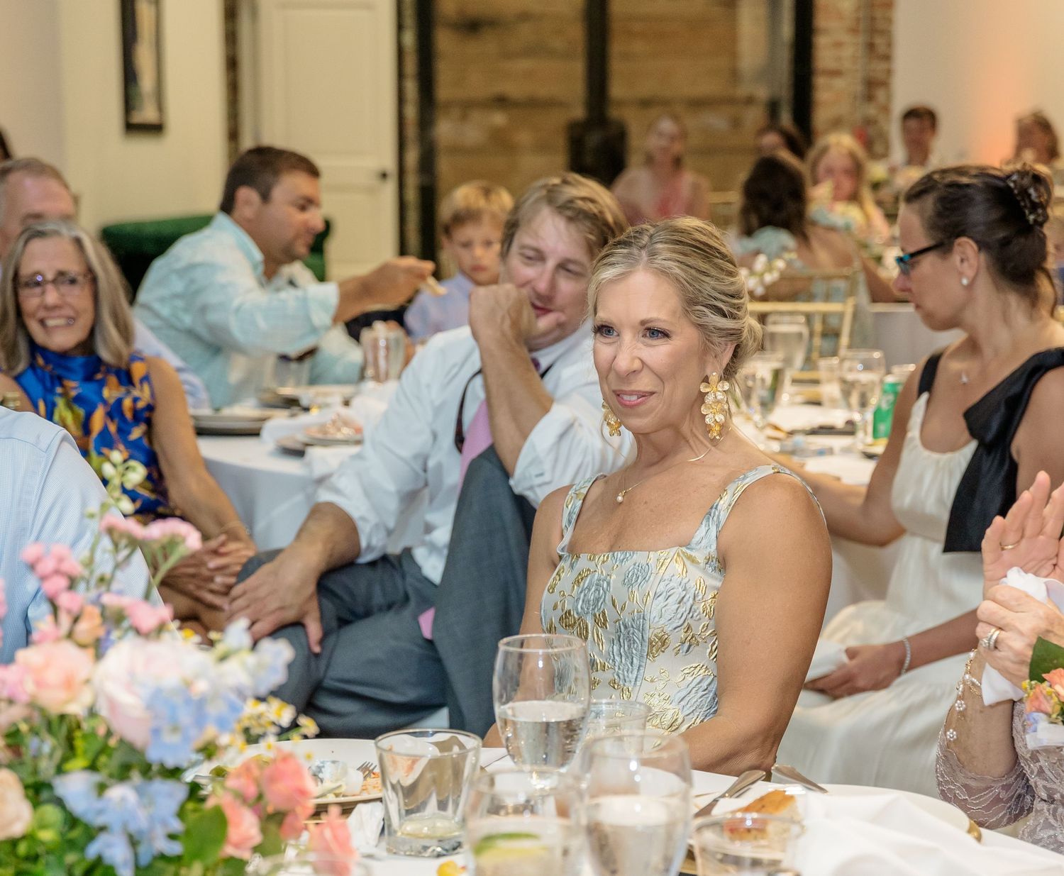 Wedding reception guests enjoy dinner and conversation at elegantly decorated tables in a rustic venue.