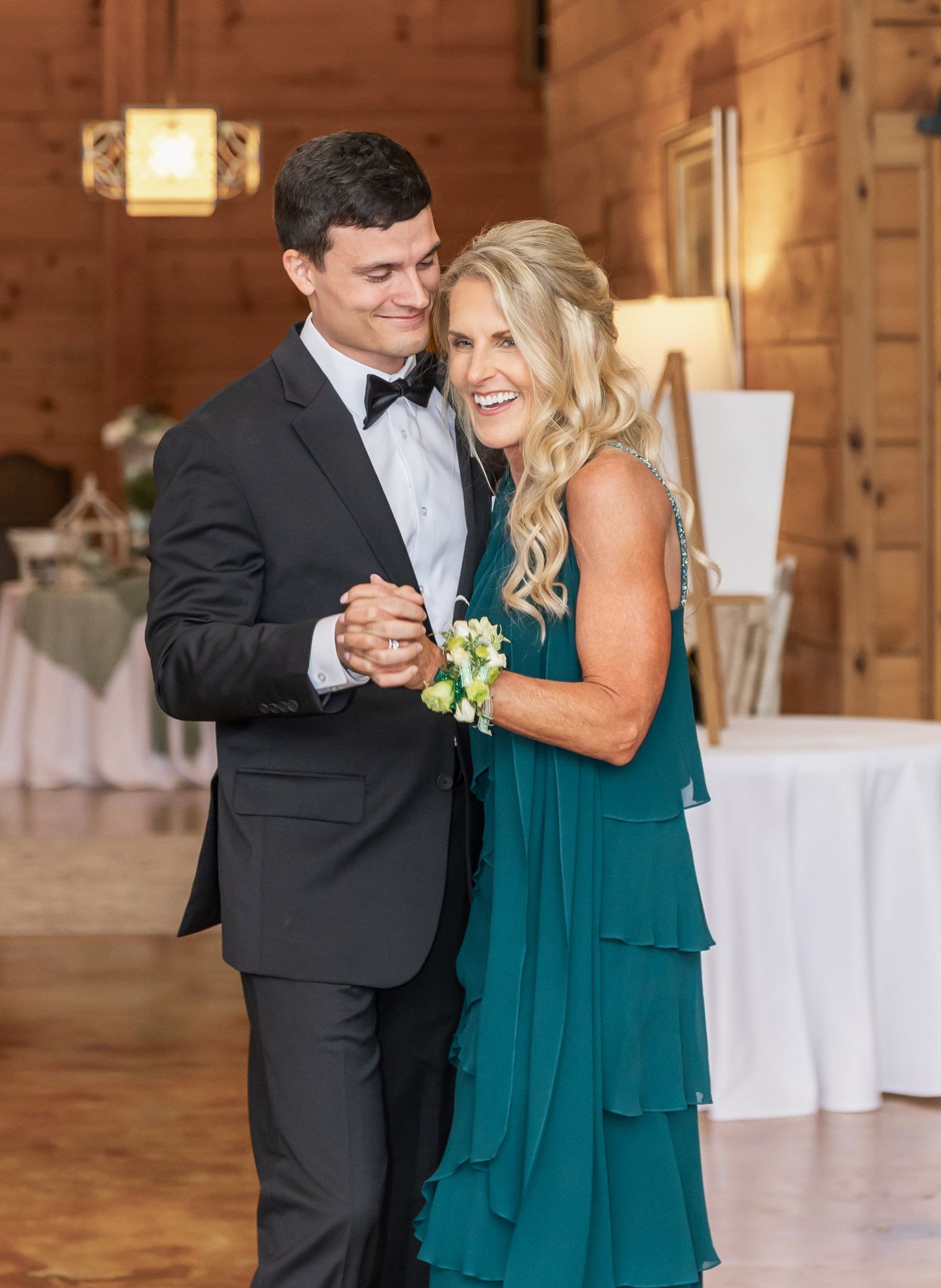 A couple dances together in a rustic barn venue, with the person in teal dress and dark suit sharing a joyful moment.