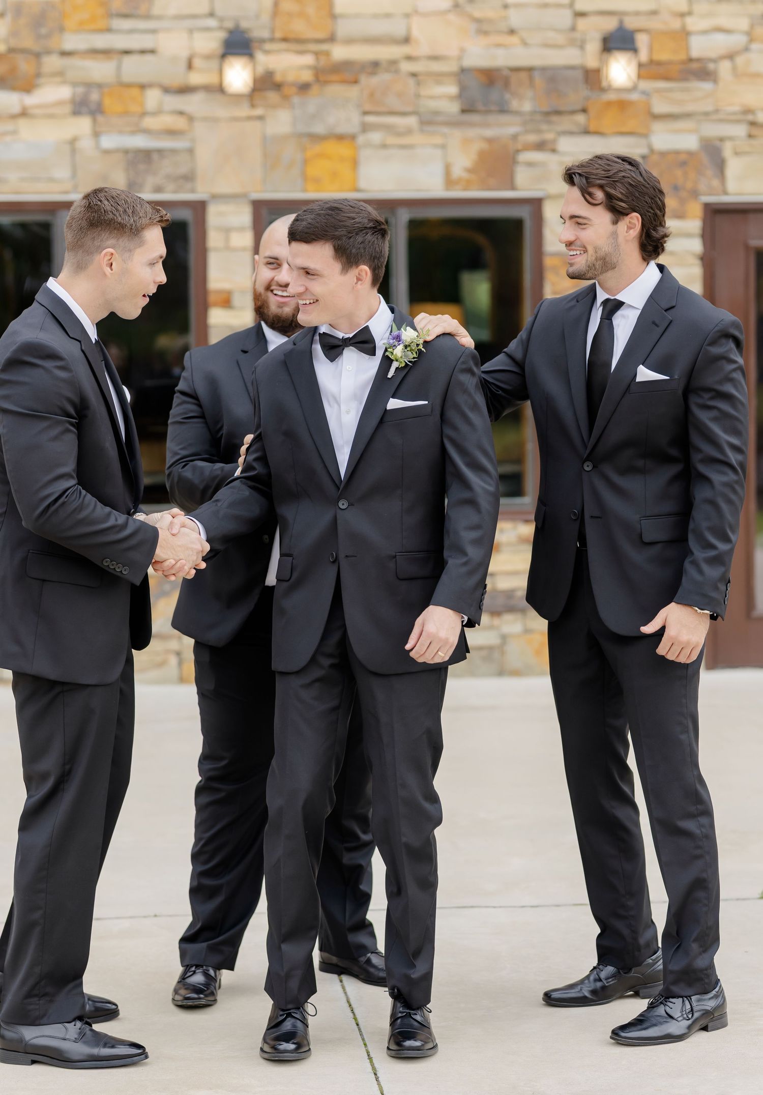 Wedding party members in black suits share a candid moment of laughter and handshakes outside a stone building.
