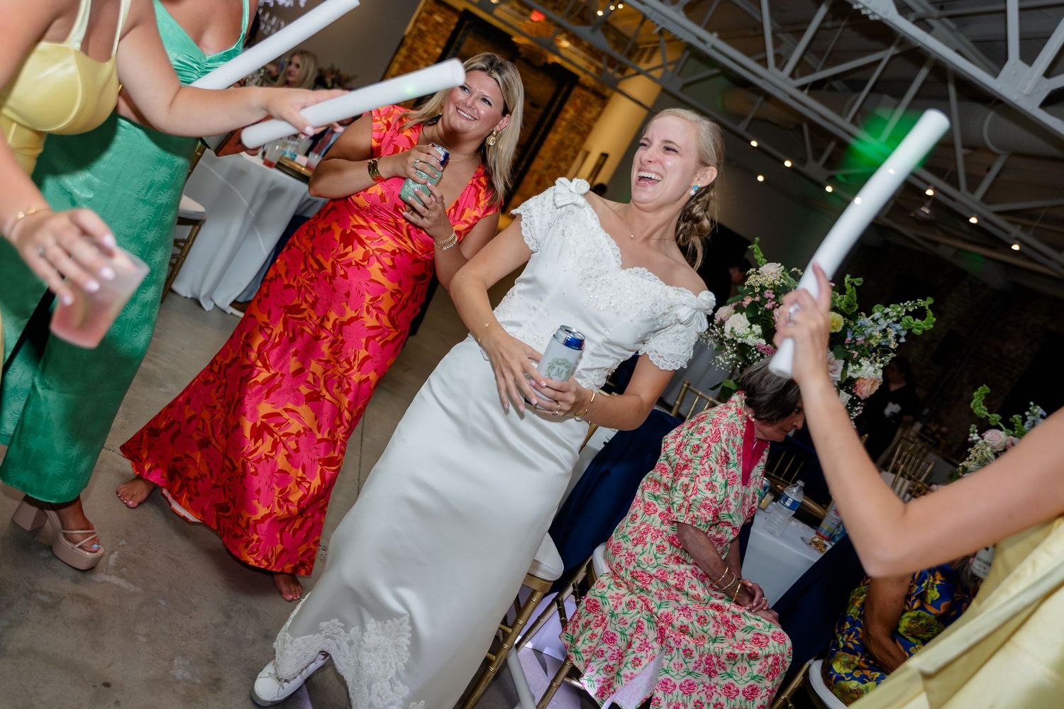 Wedding party guests dance with glowing light sticks at an indoor reception celebration.