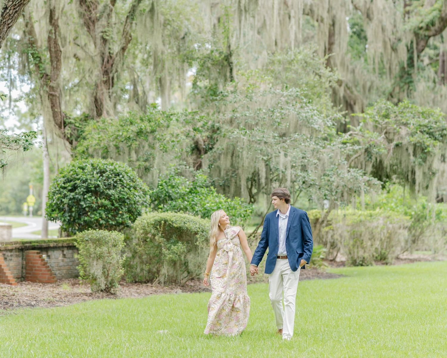 A couple walks hand in hand through a scenic garden with Spanish moss hanging from tall trees.