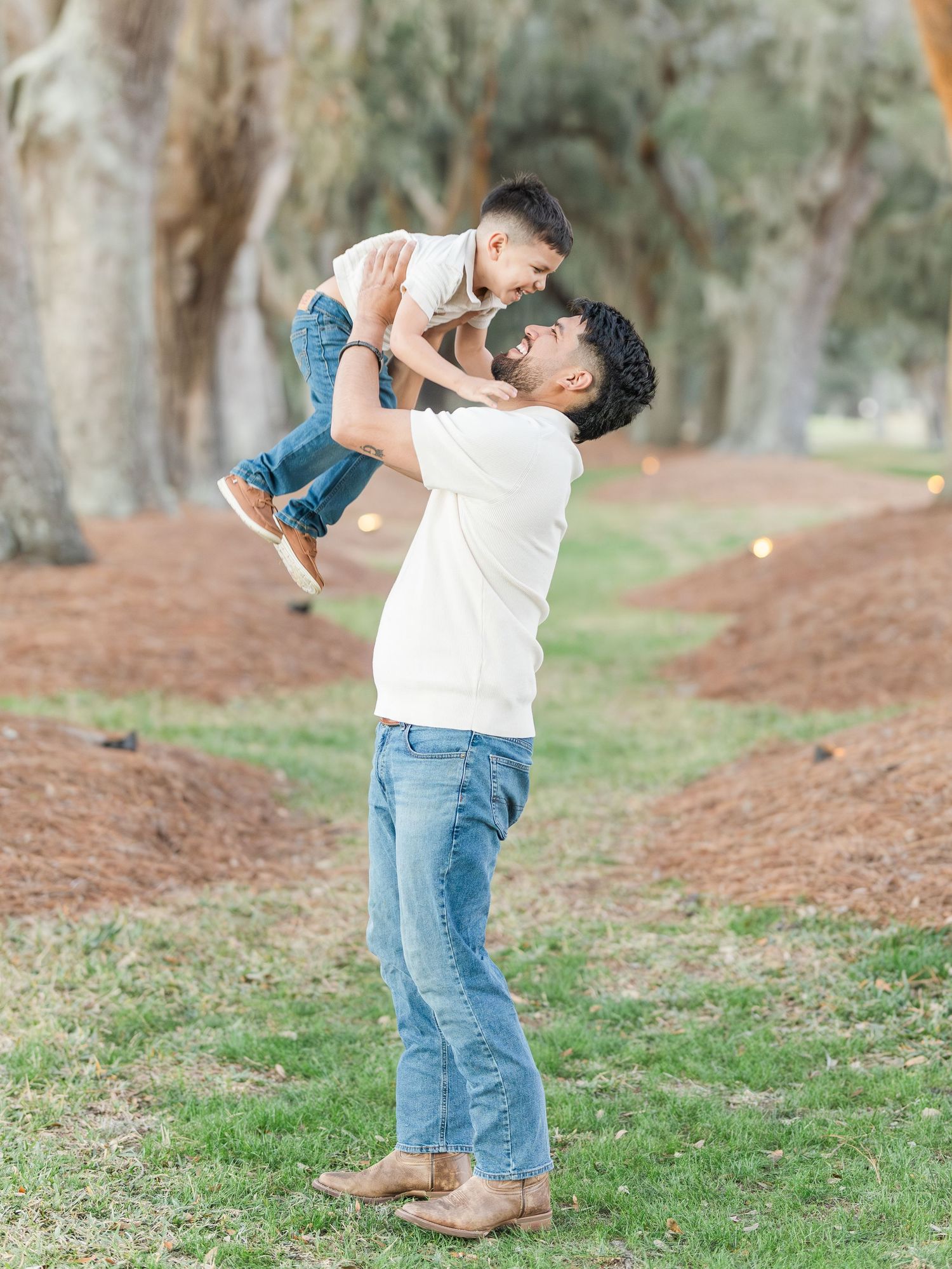 Parent playing and lifting child in air against backdrop of tall trees.