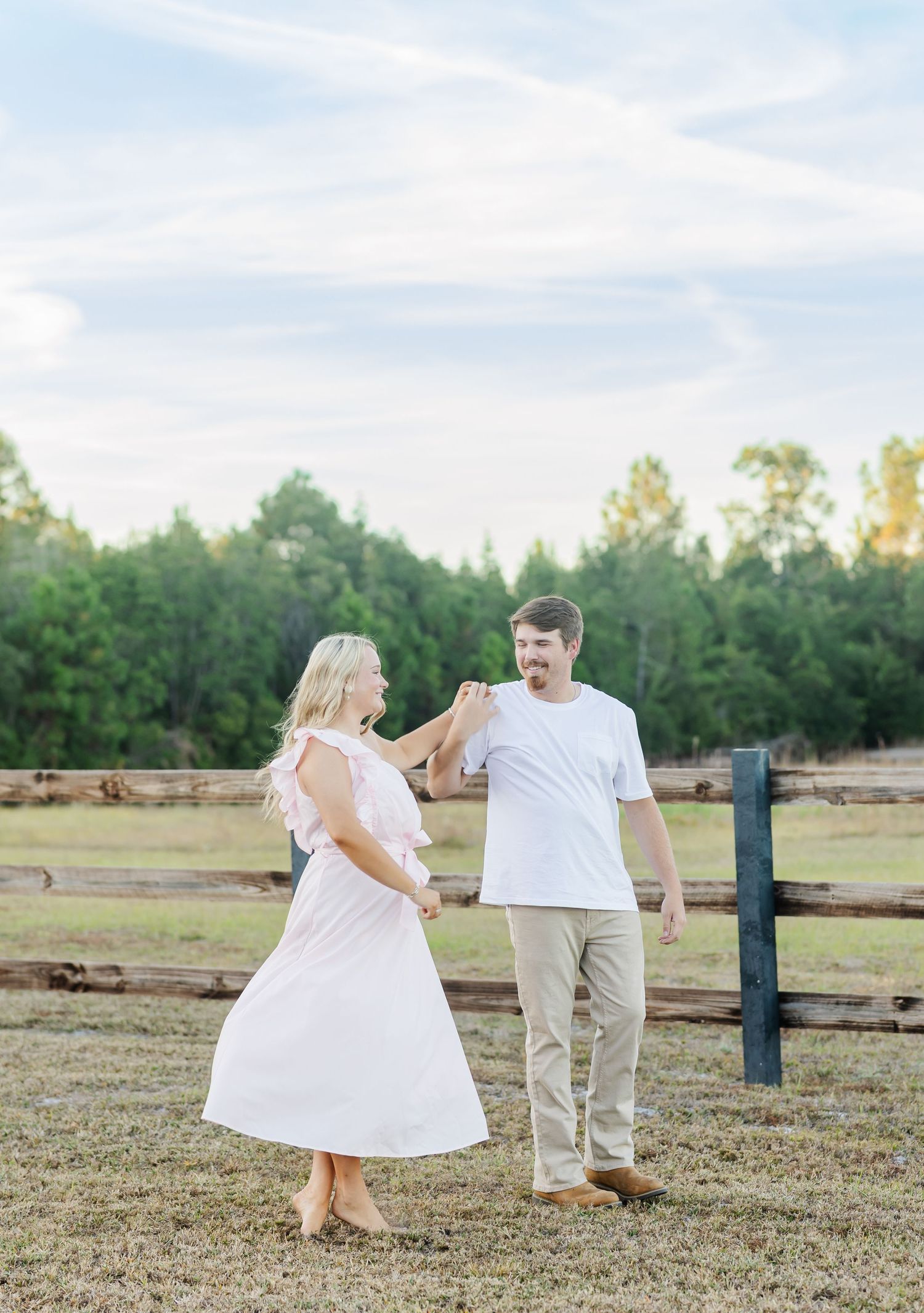 A couple dances playfully in a grassy field at sunset with trees in the background.