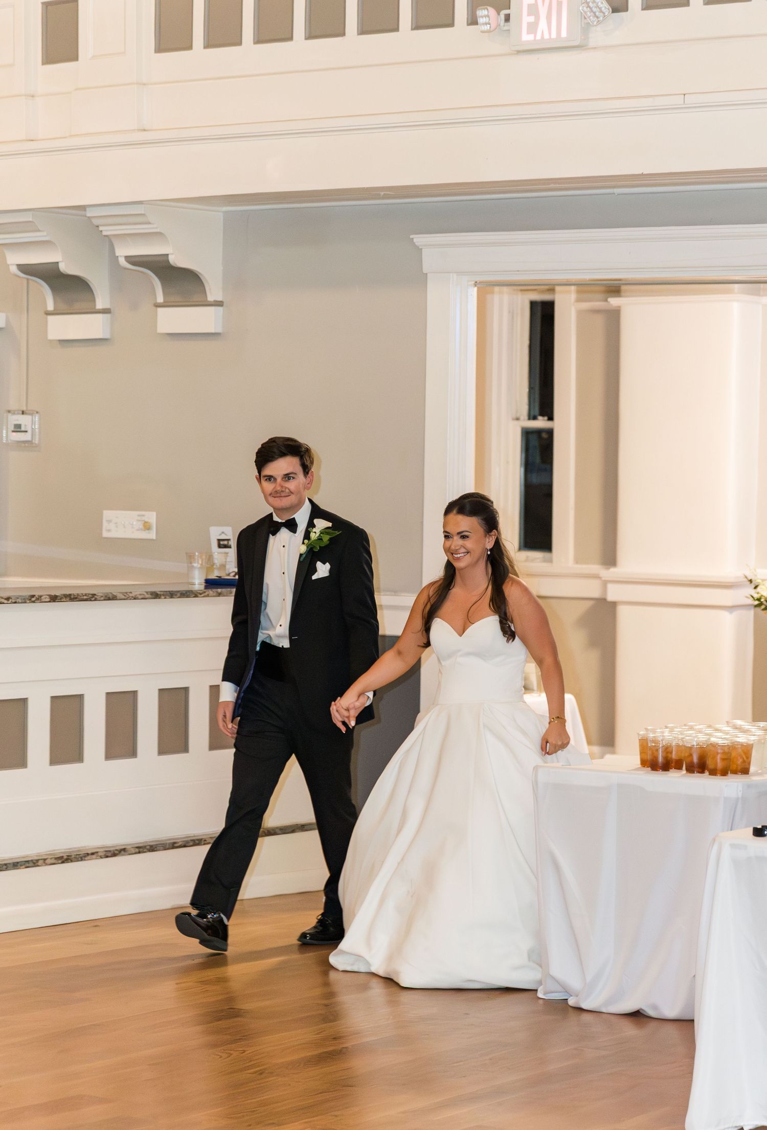 Bride and groom hold hands walking into their wedding reception, she in a white gown, he in a black tuxedo.