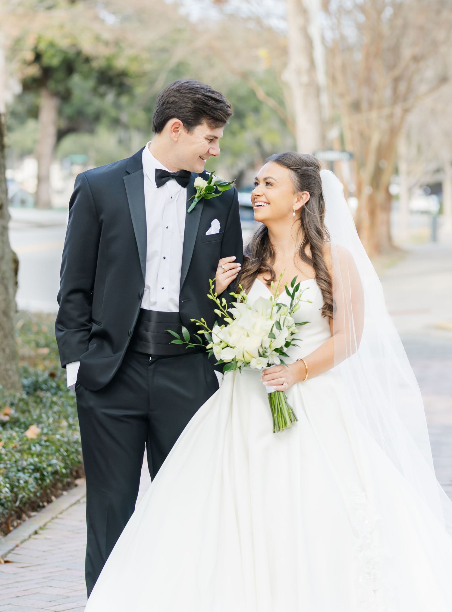 Bride in white ballgown with veil holds white bouquet beside groom in black tuxedo on tree-lined sidewalk.