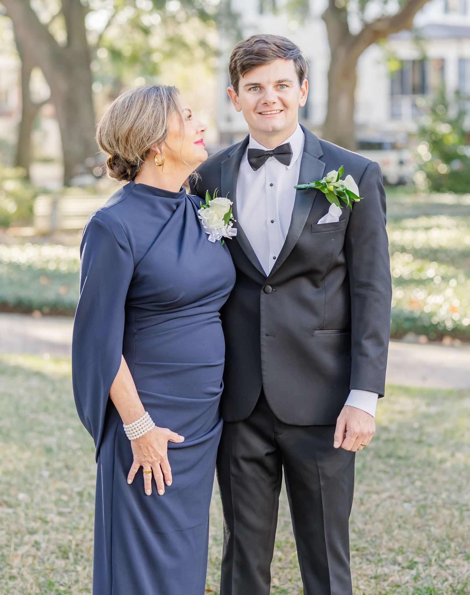 Groom in black tuxedo with bow tie stands beside woman in navy blue gown at outdoor wedding.