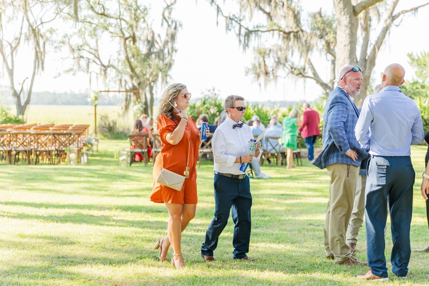 Guests mingle on a sunny lawn with wooden chairs and Spanish moss draped trees in the background at an outdoor event.