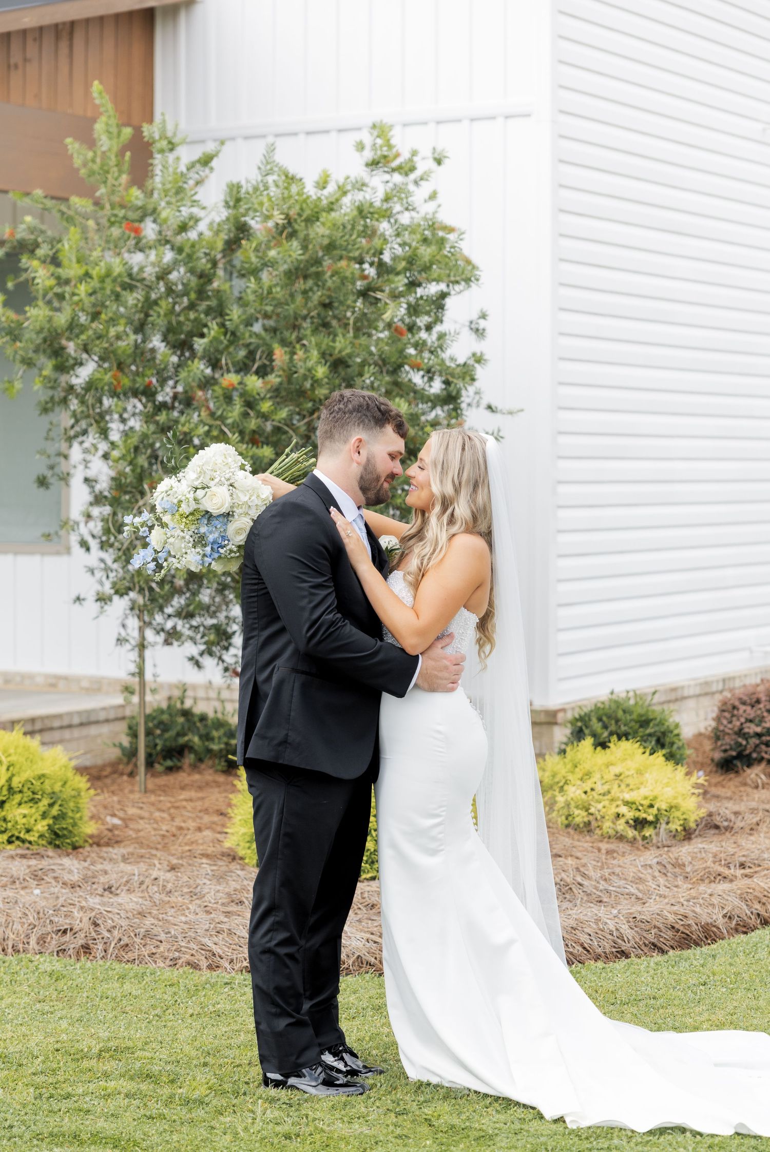 Bride and groom sharing a romantic kiss outdoors, bride in white gown with veil holding blue hydrangea bouquet.