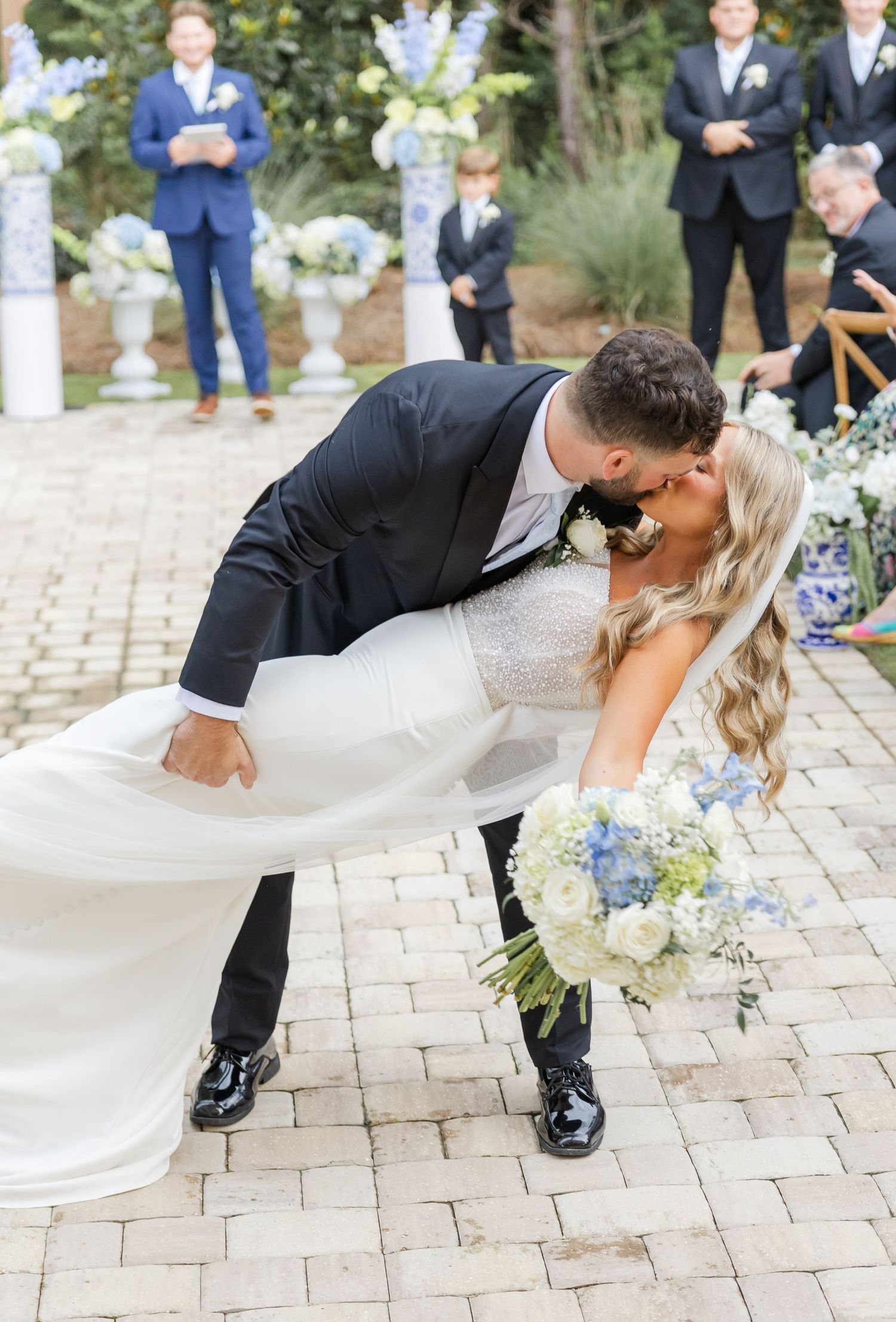 Groom dips bride for a kiss at outdoor wedding ceremony, bride holding blue and white floral bouquet in white gown.