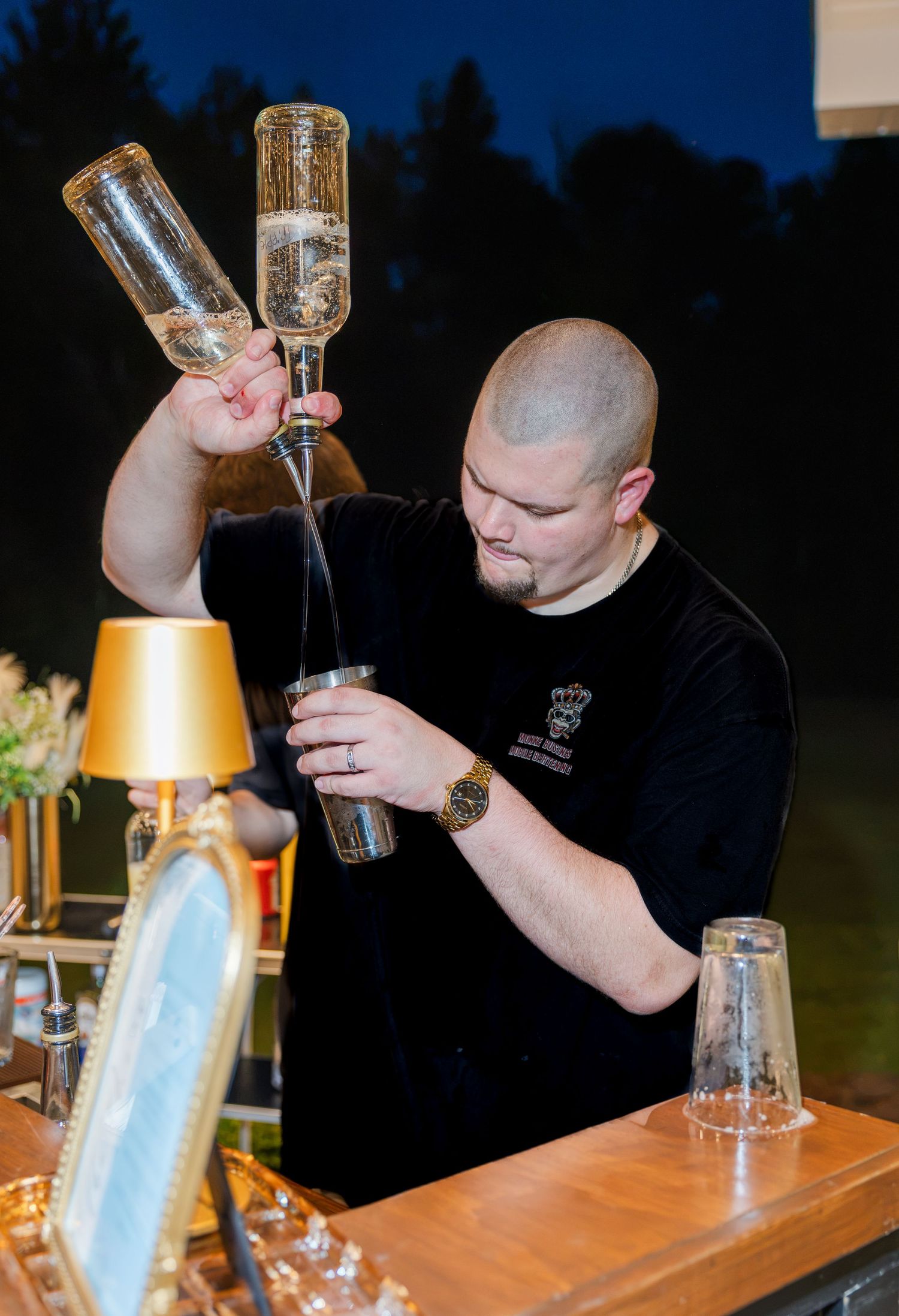 Bartender pouring champagne into flutes at an outdoor evening wedding bar with gold accents and elegant decor.