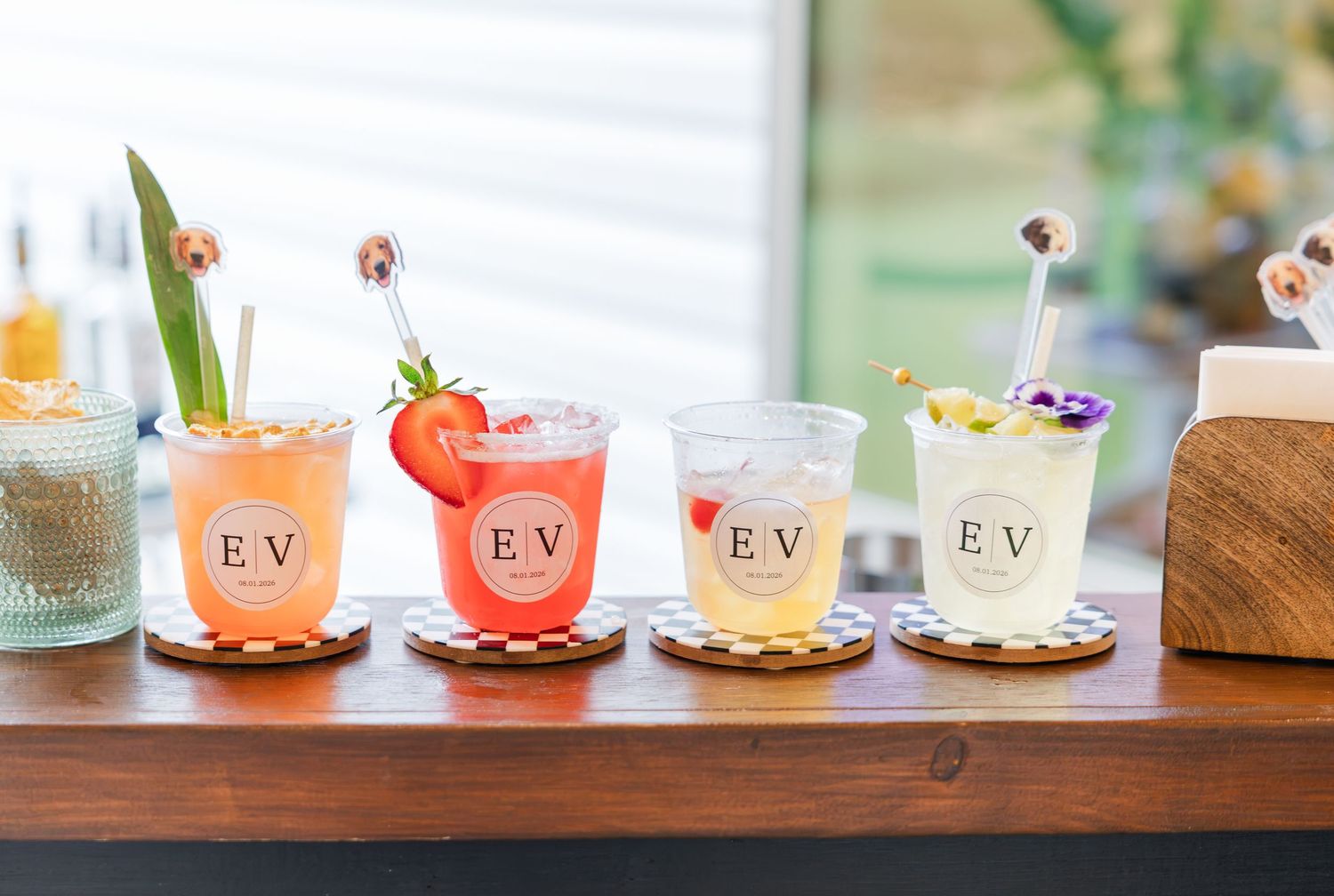Four signature cocktails in monogrammed EV cups displayed on a wooden bar at a wedding reception.