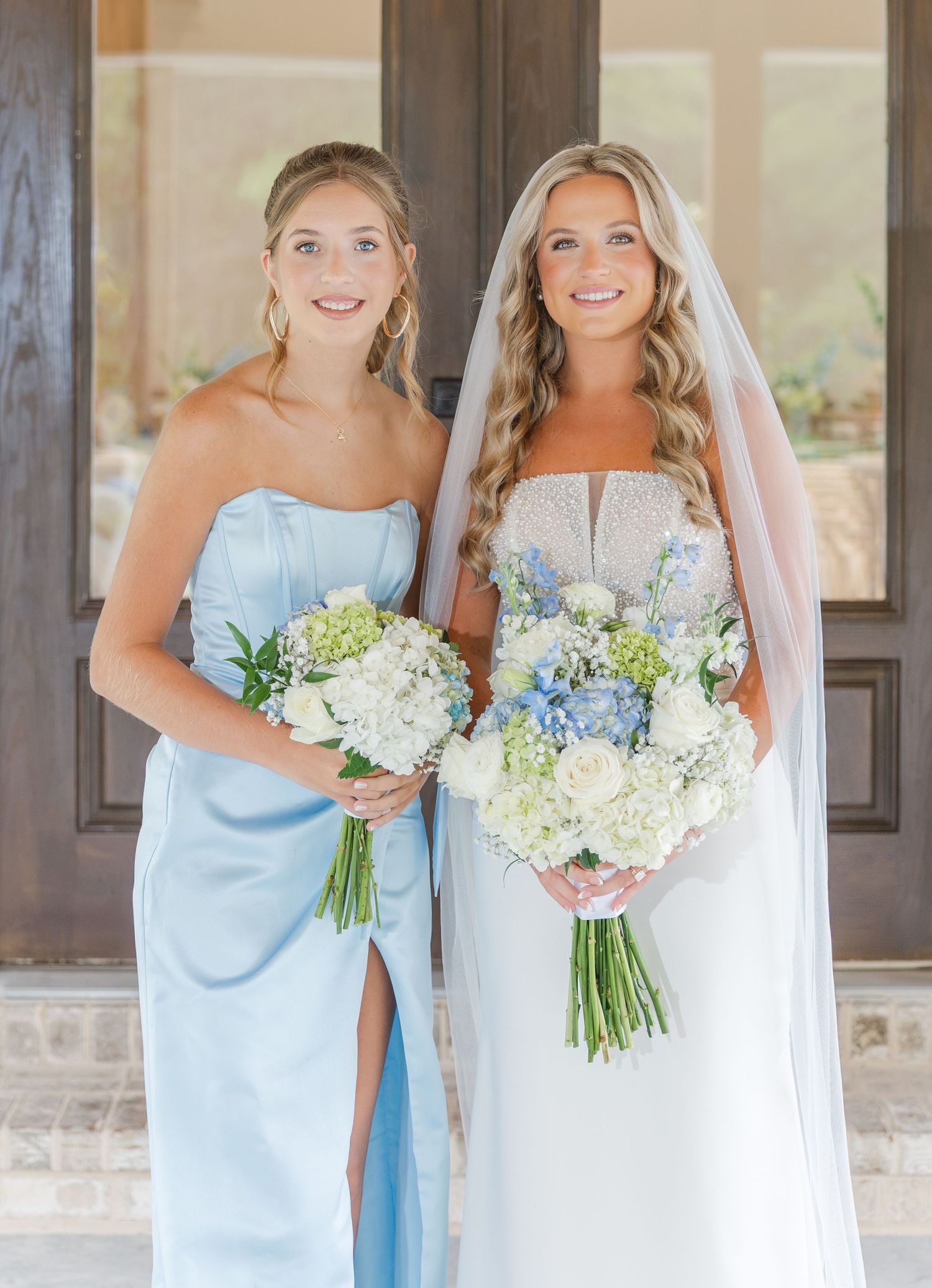 Bride in pearl wedding gown with veil and bridesmaid in strapless light blue gown holding matching white and blue bouquets.