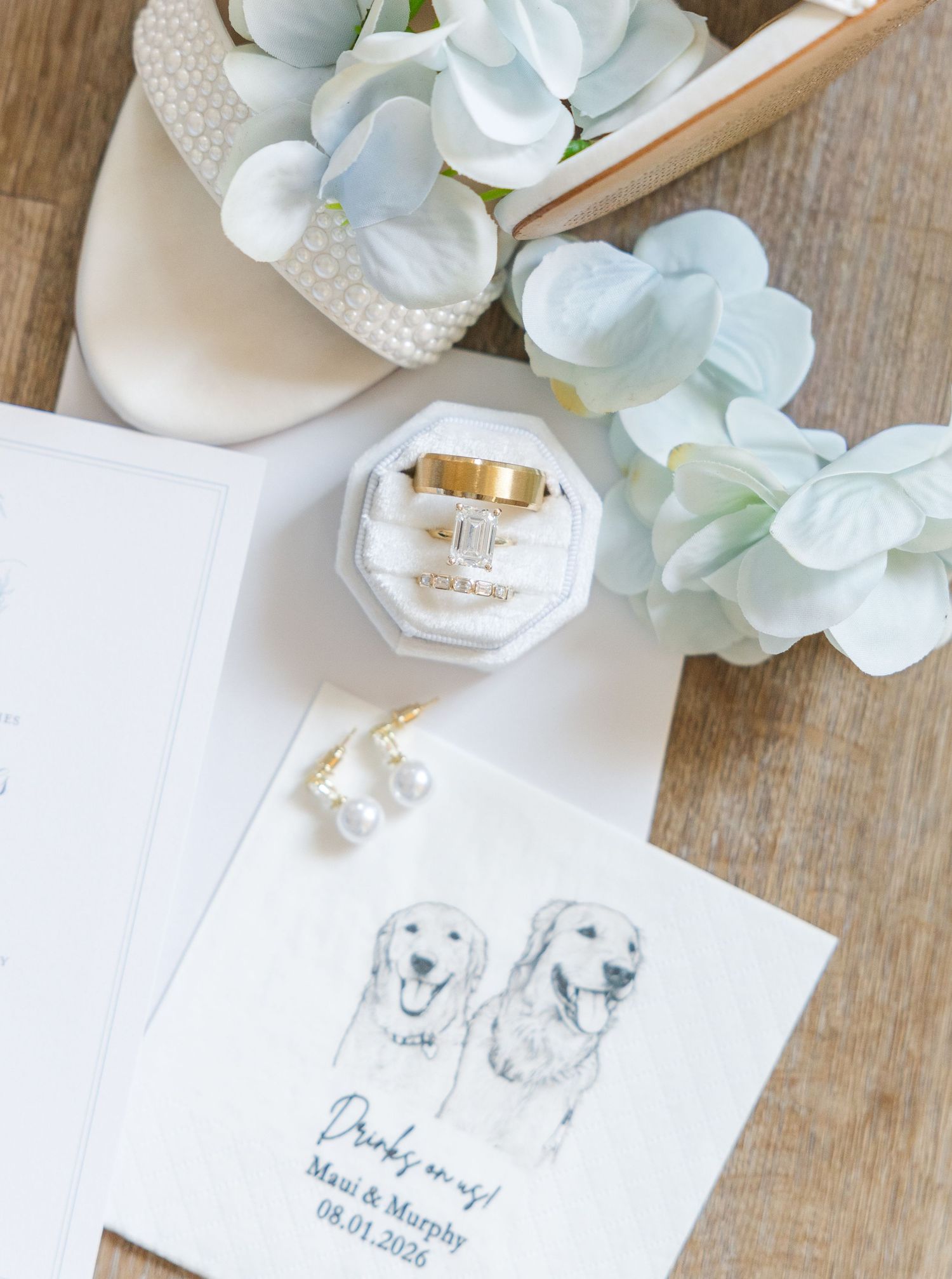 Wedding flat lay with gold rings in octagonal box, pearl earrings, custom dog napkins, bridal shoes, and blue hydrangeas.