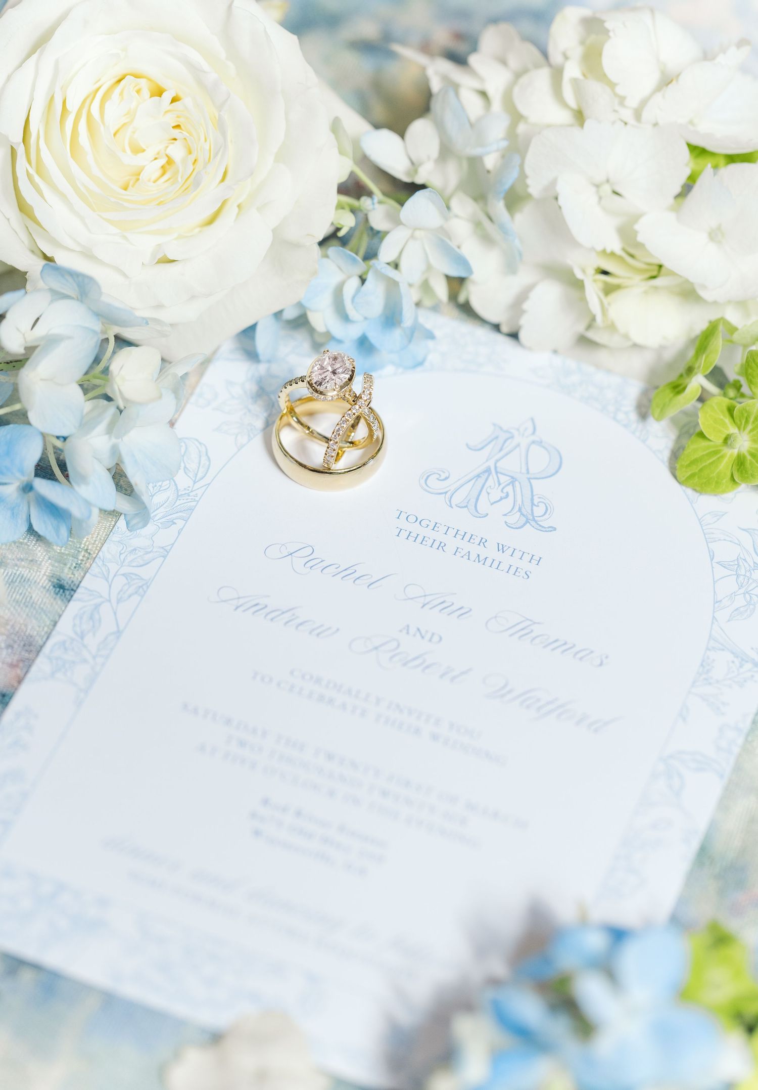 Wedding invitation with blue monogram design surrounded by white roses and hydrangeas with gold engagement ring on top.