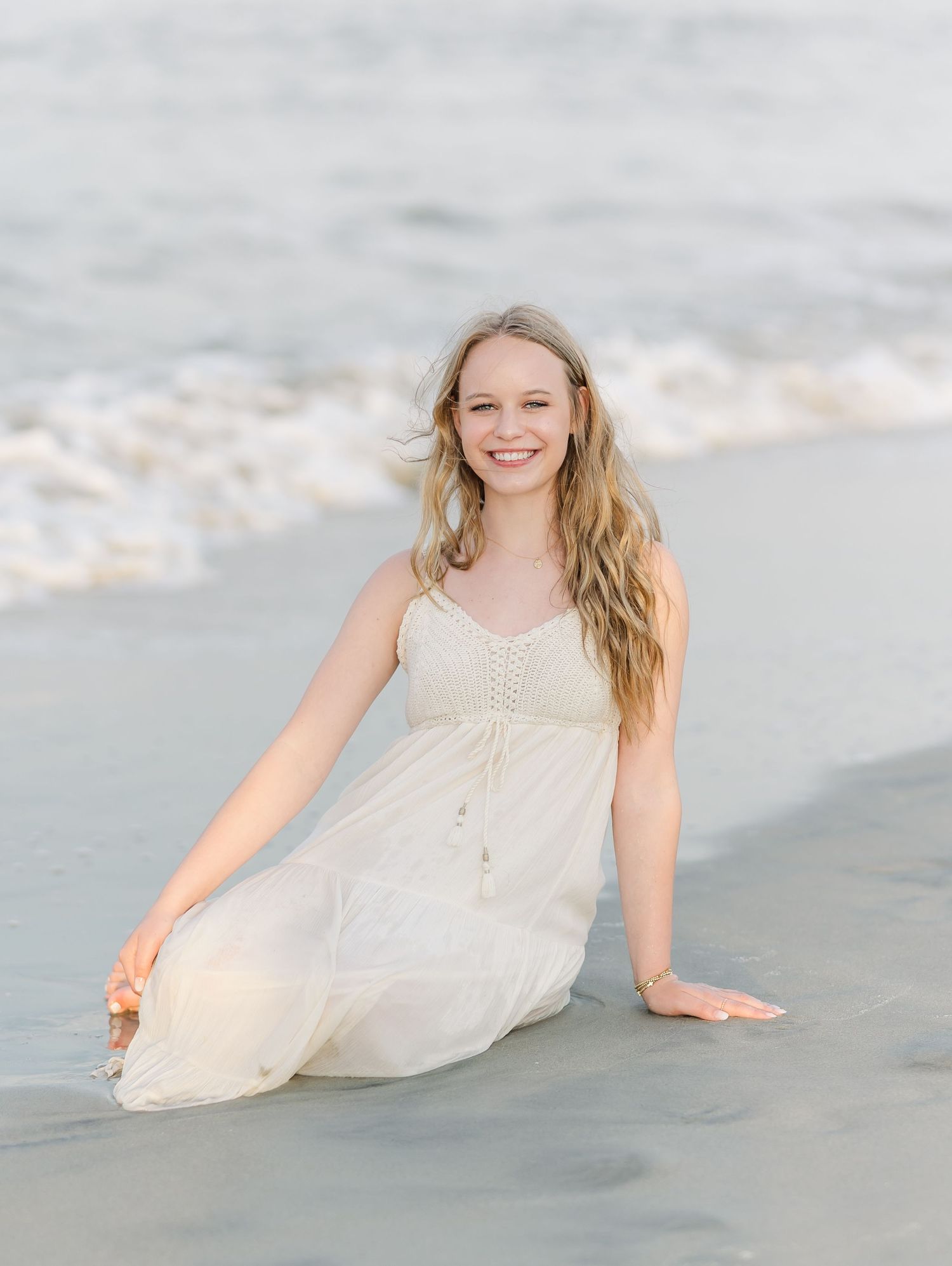 A person in a flowy white dress sits on the beach with waves in the background during golden hour.