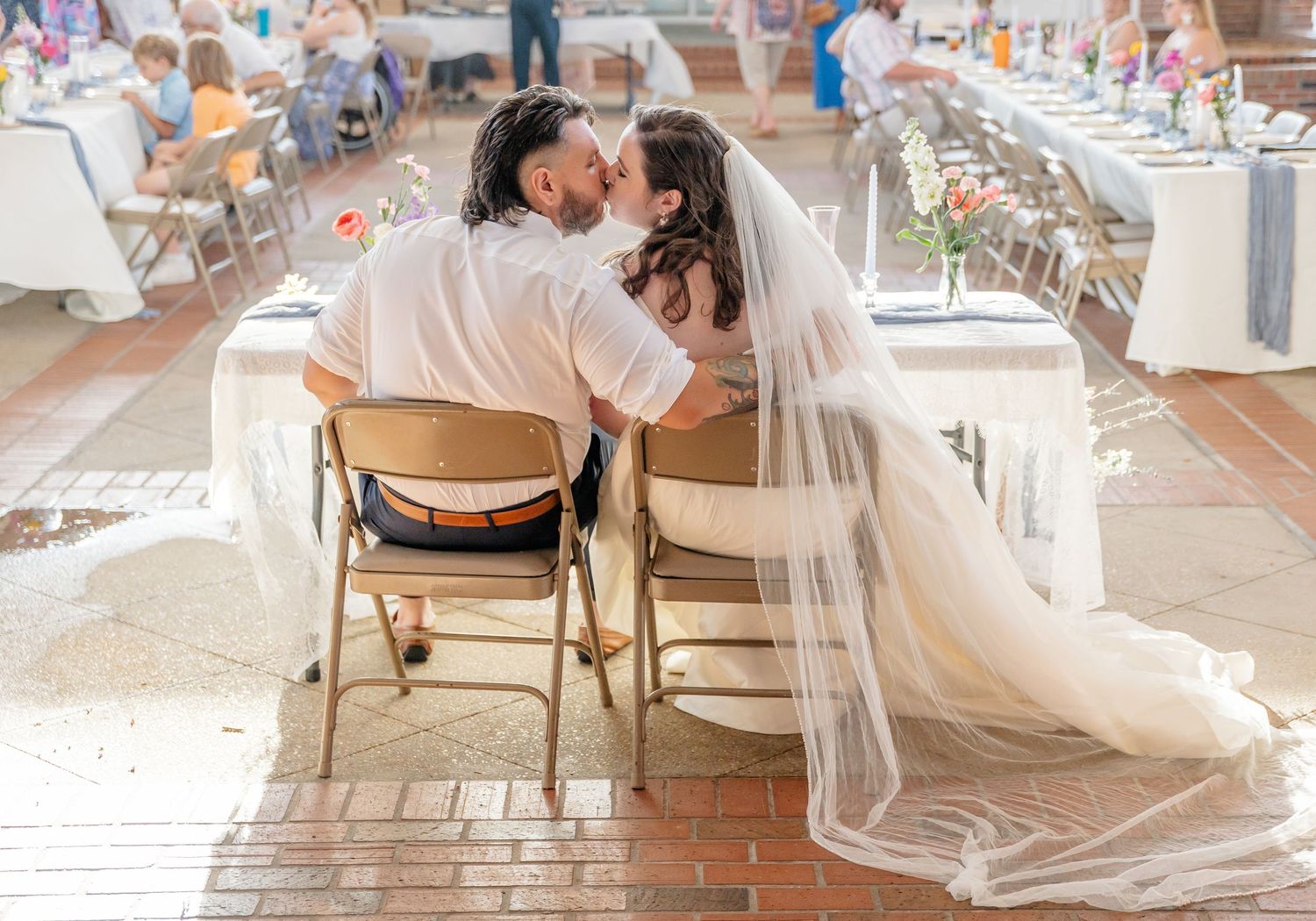 A couple in wedding attire share an intimate moment seated in folding chairs at their reception venue.