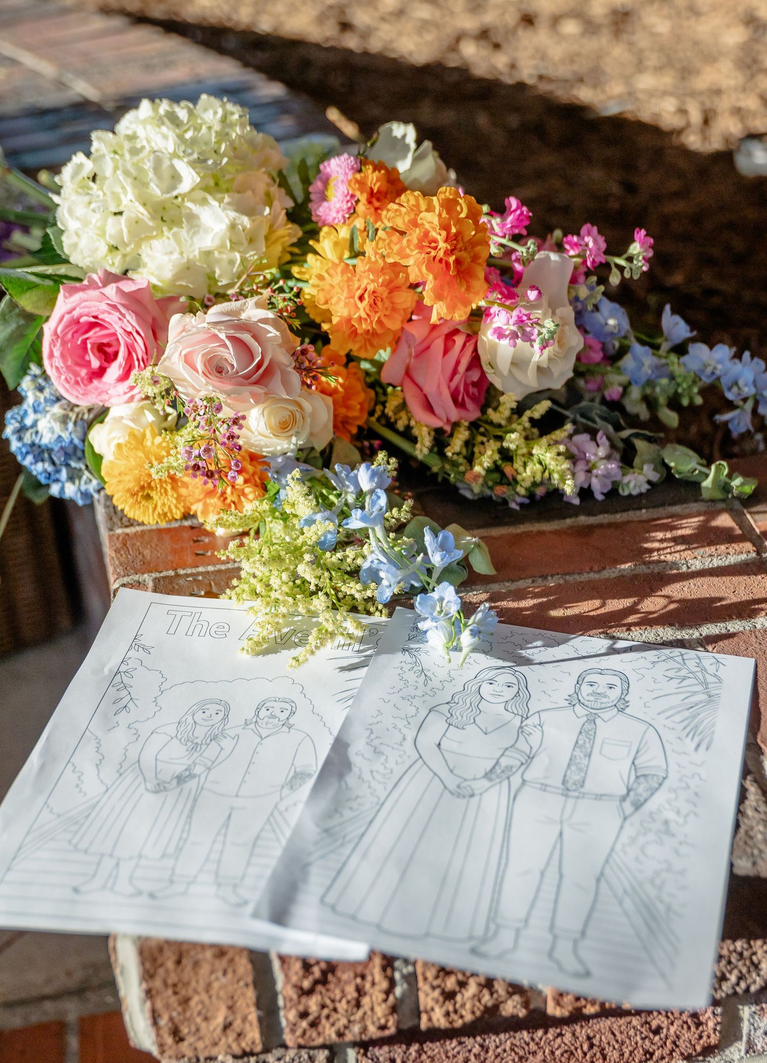 A bouquet of pink roses, orange marigolds, blue hydrangeas and white flowers sits next to sketches on a wooden surface.