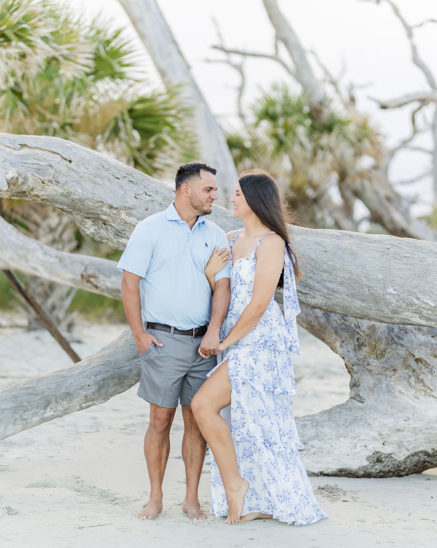 A couple in coordinating blue and white outfits share a romantic moment on a beach with driftwood and palm trees.