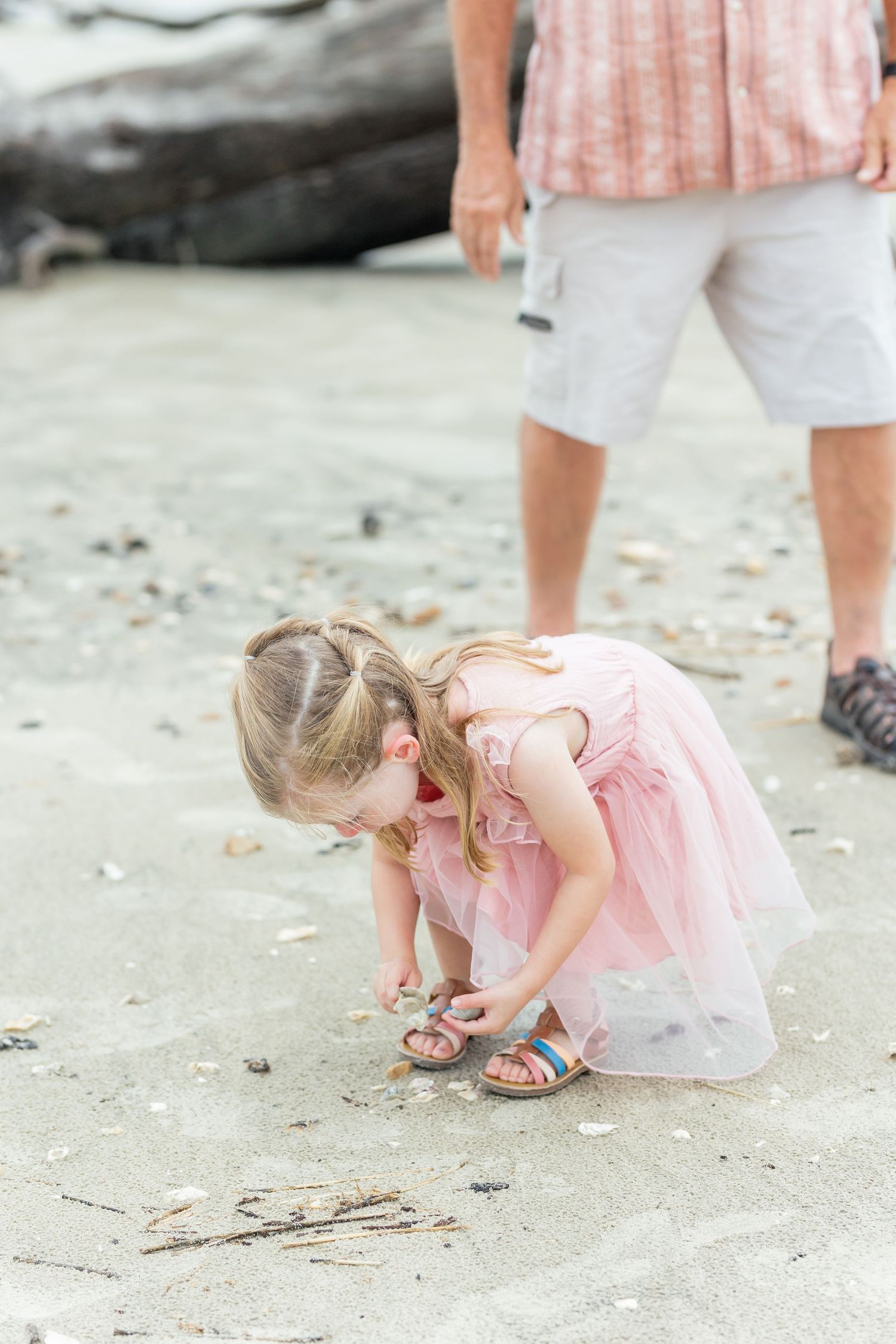 A small child in a pink tulle dress bends down to examine something on a sandy beach while an adult stands nearby.