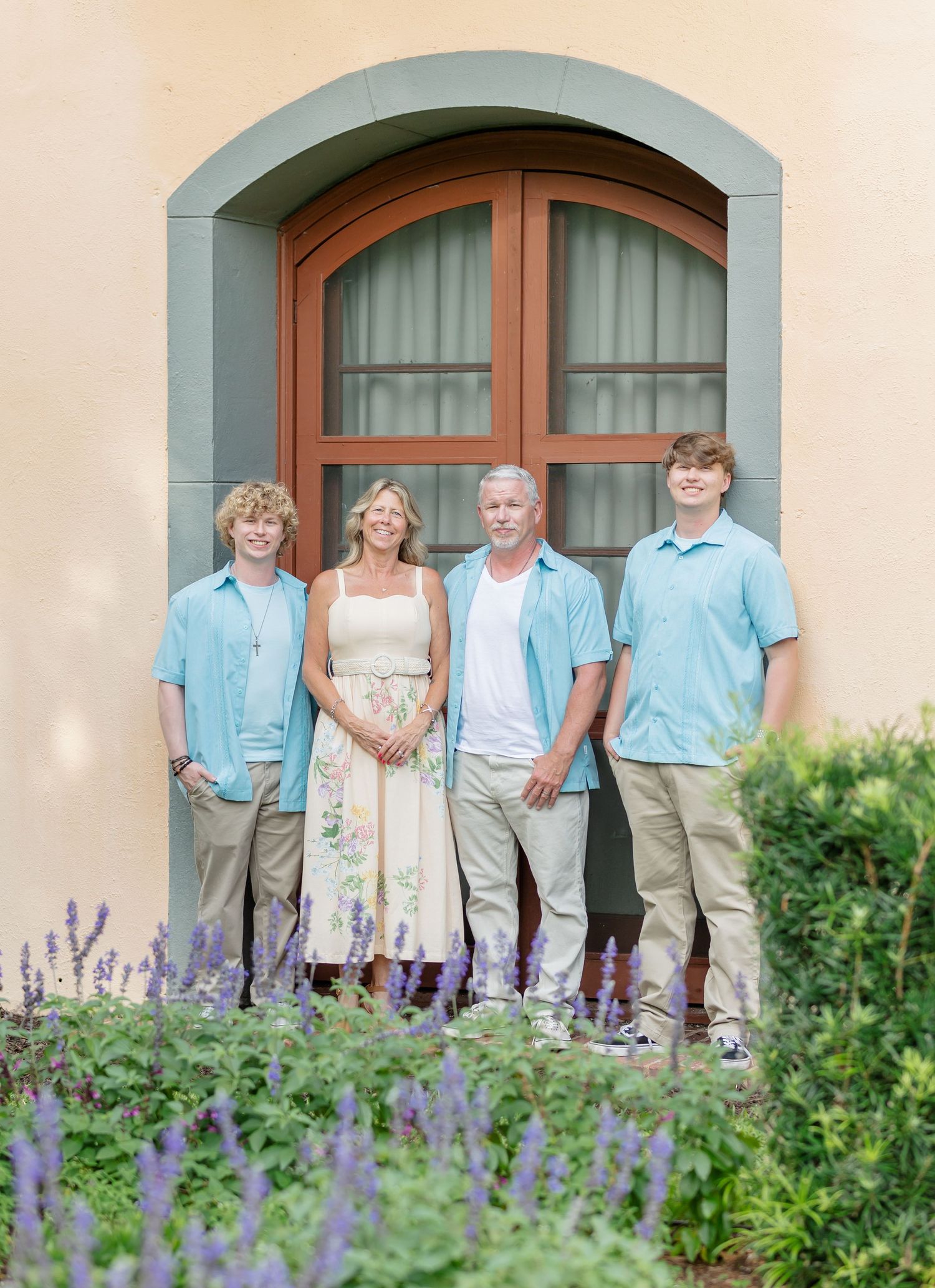 Four people in coordinating light blue and white outfits pose in front of an arched doorway with lavender flowers in foreground.