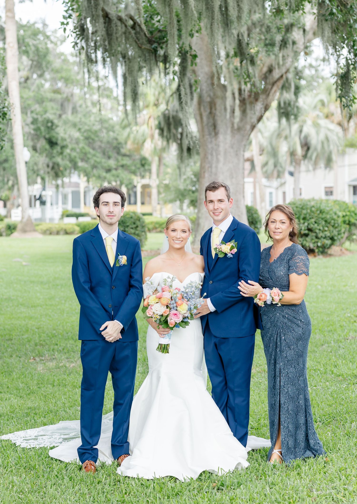 Wedding party portrait featuring formal attire in navy and gray tones under moss-draped trees.