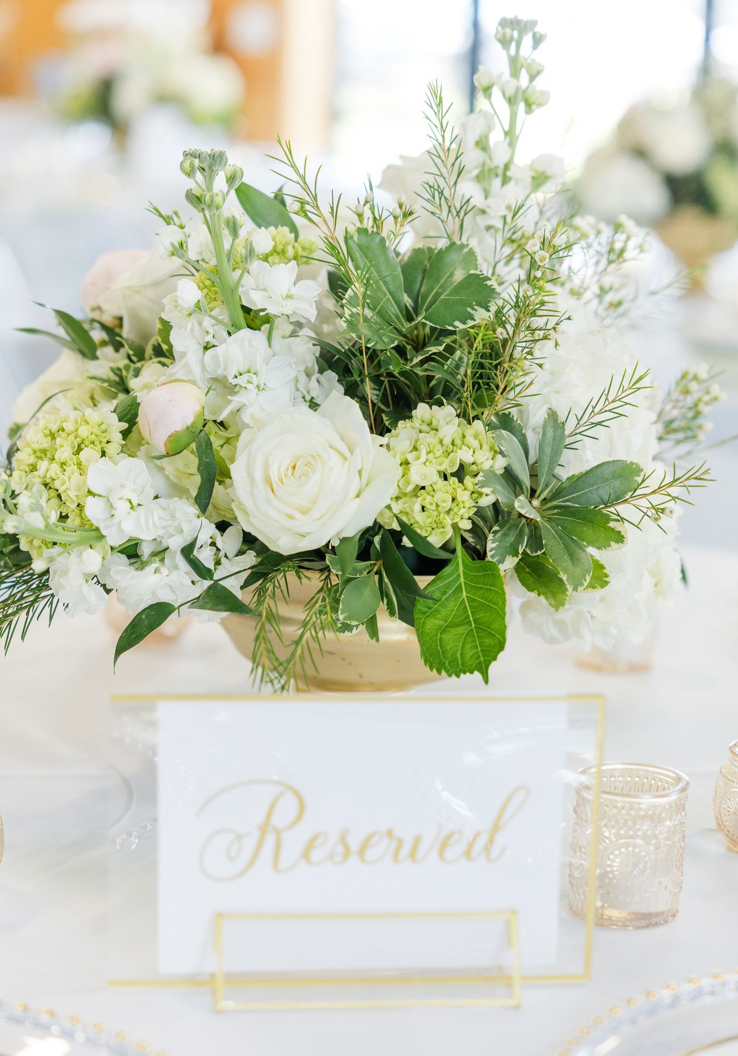 Elegant wedding table centerpiece with white roses, hydrangeas, and greenery beside a gold-framed Reserved sign.