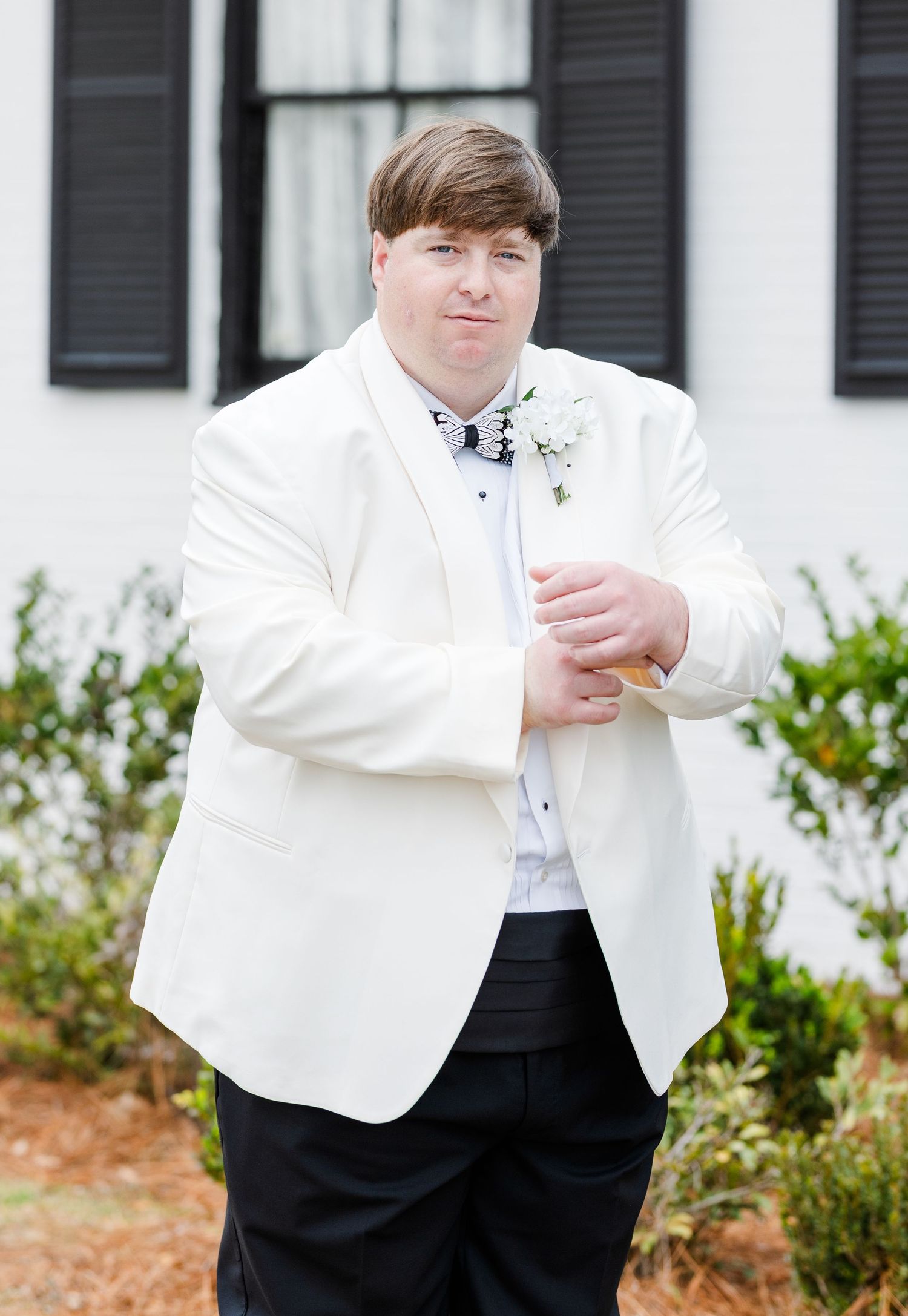 Groom in a white tuxedo jacket with a floral boutonniere posing outside a white building with black shutters.