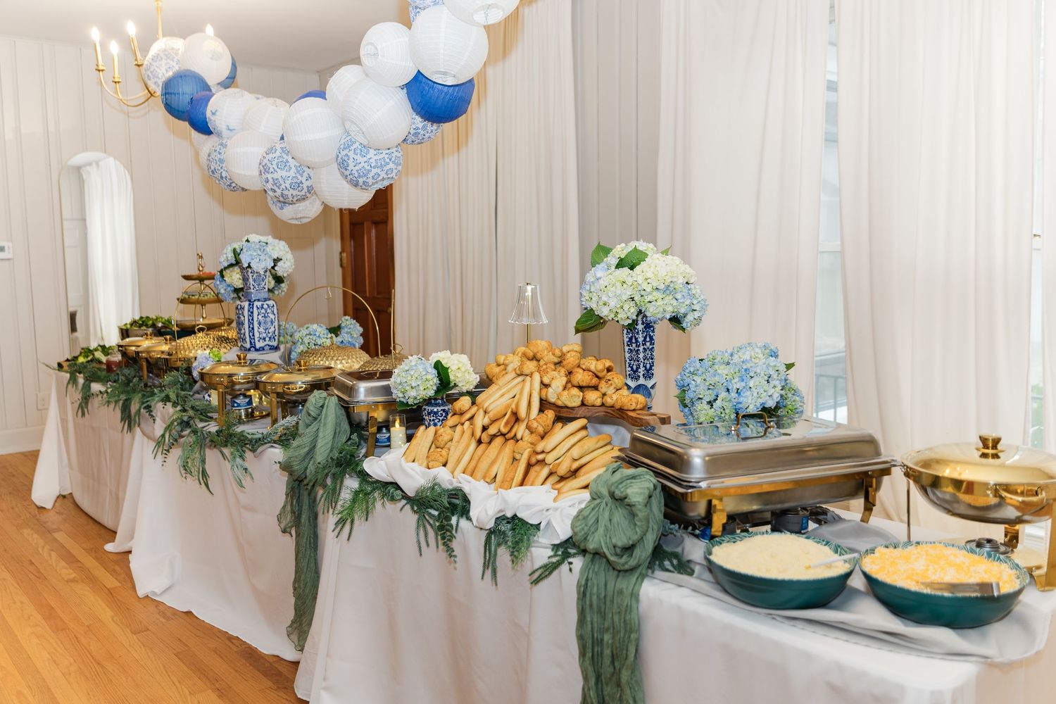 Elegant wedding reception buffet table with blue hydrangeas, balloon arch, and catered food in chafing dishes.