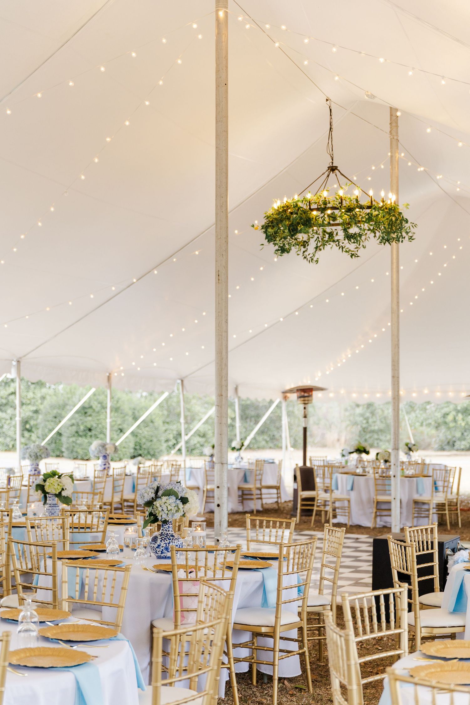 Elegant white wedding tent reception with string lights, greenery chandelier, gold chiavari chairs, and blue table decor.