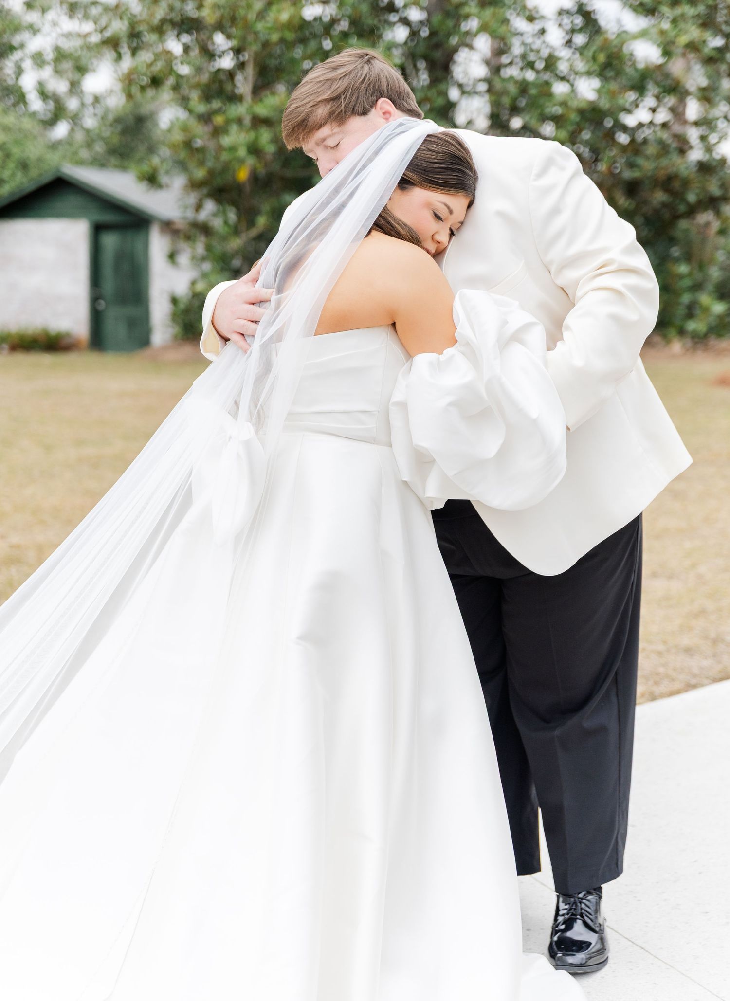 Romantic first look moment as groom in white tuxedo embraces bride in strapless gown with flowing cathedral veil.