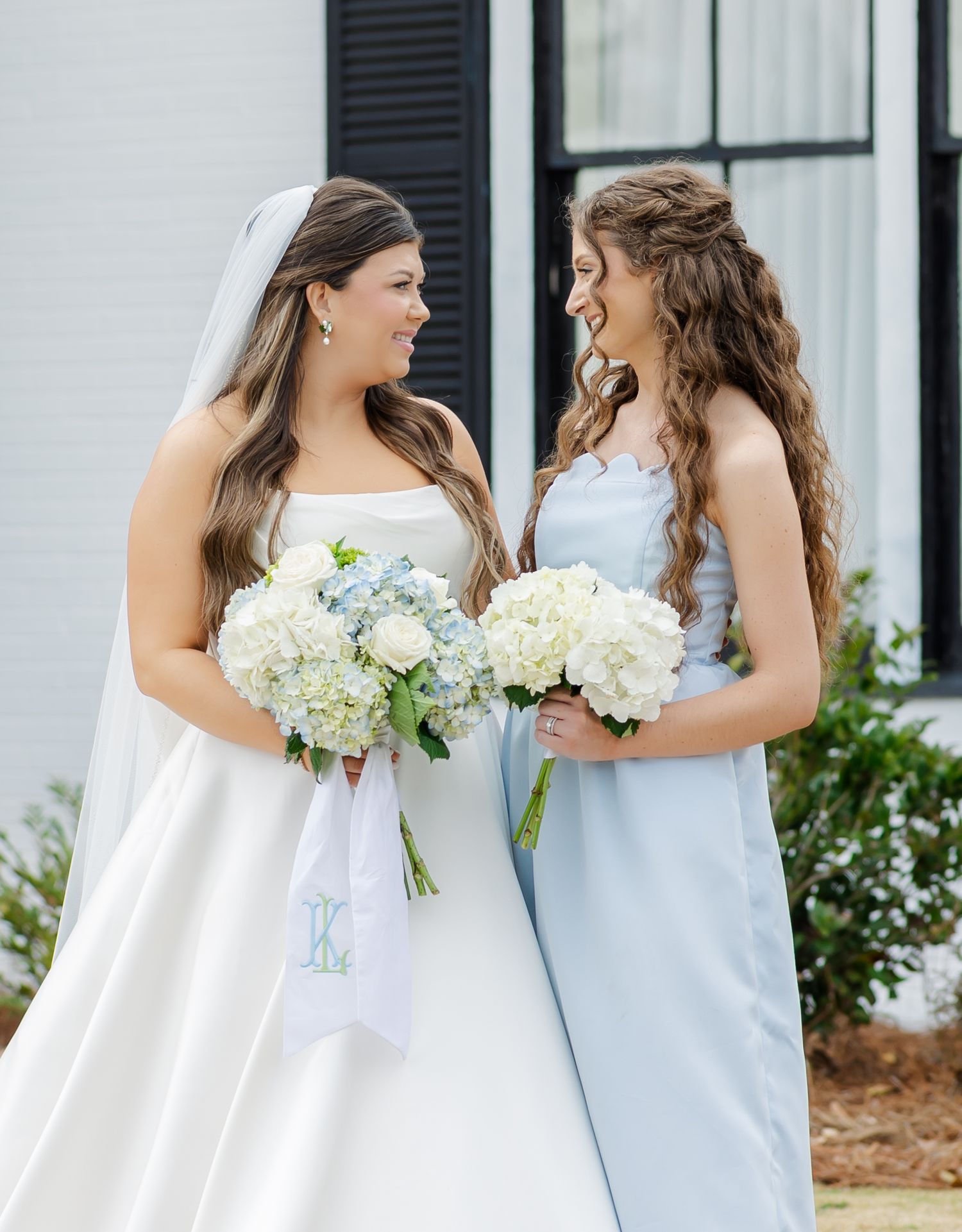 Bride in white strapless gown and bridesmaid in light blue dress holding white hydrangea bouquets smiling at each other.