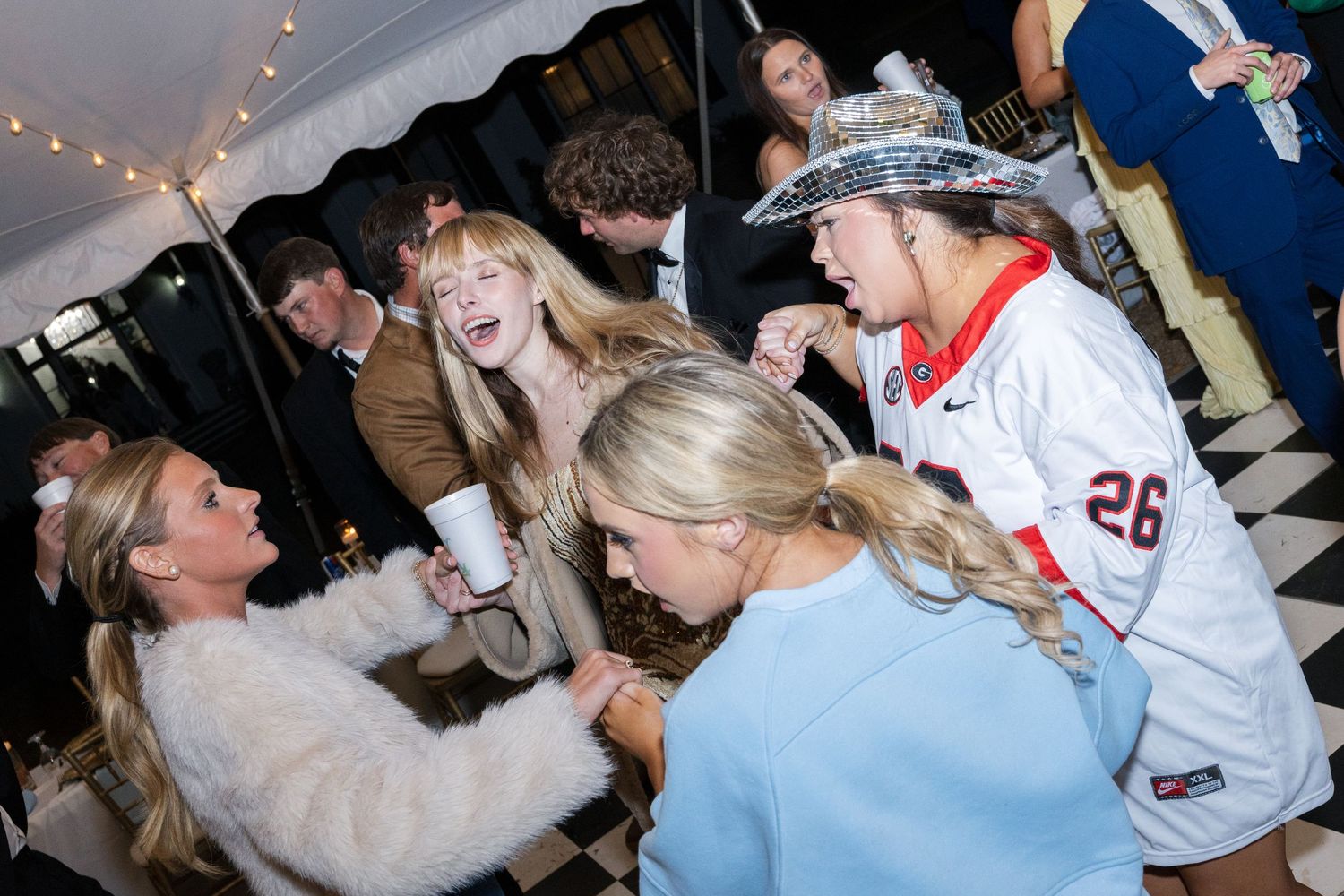 Wedding guests laugh and dance together at a lively evening reception under a white tent.