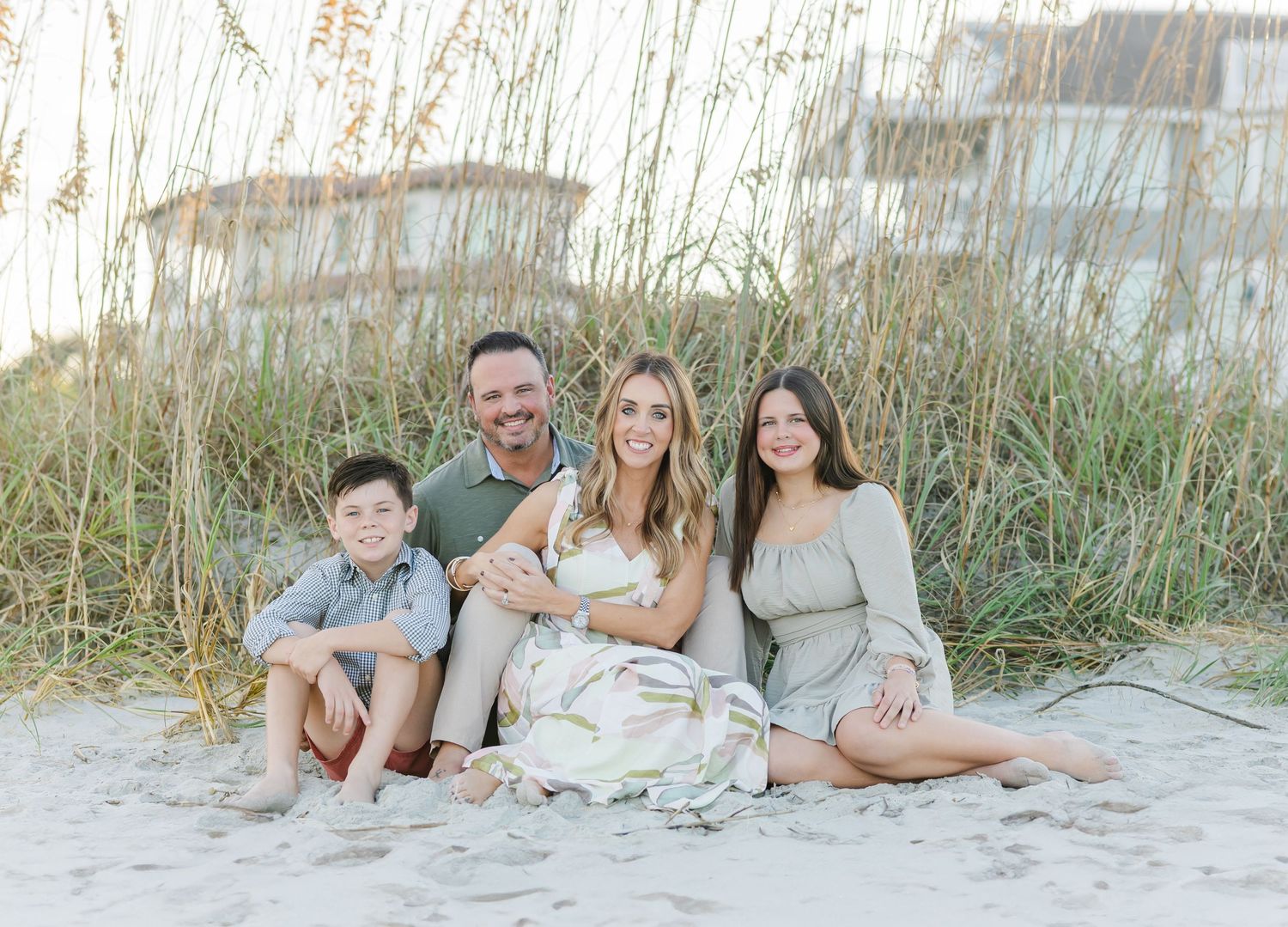 Family group lounging together on sandy beach at sunset with sea grass in background.