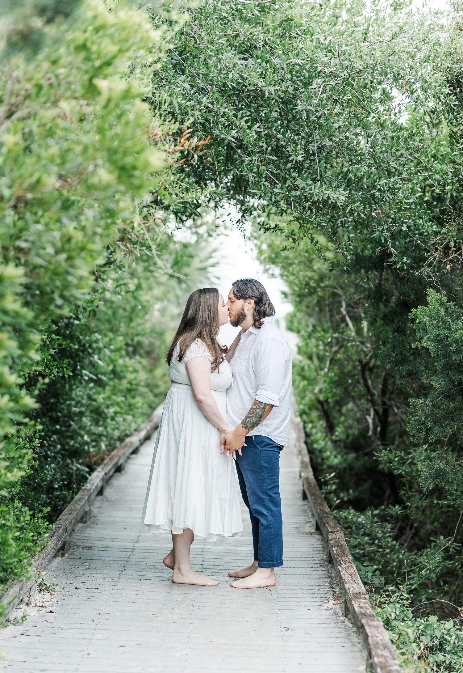 Couple shares an intimate moment on a tree-lined boardwalk in white and blue outfits.