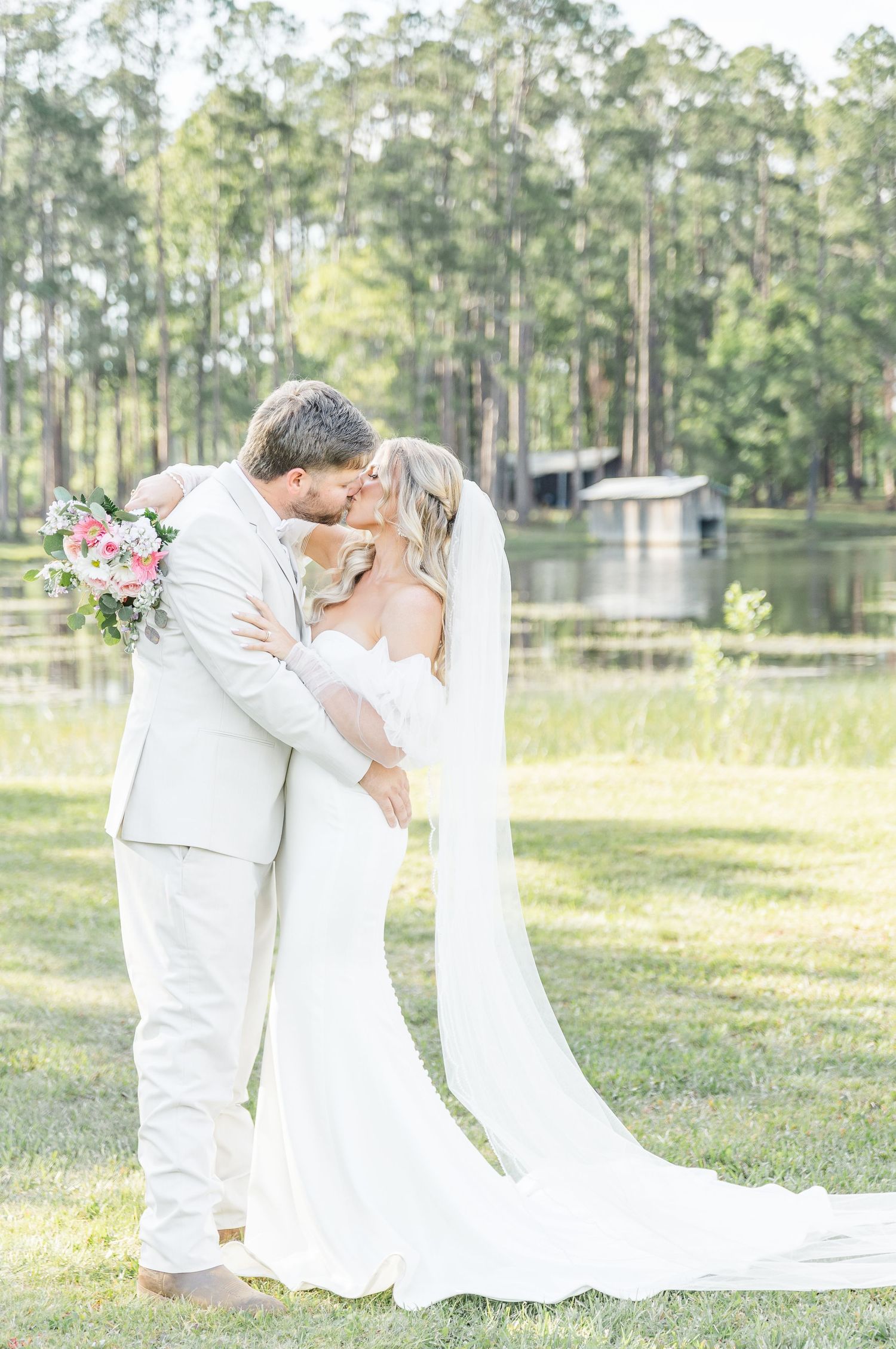 A couple in all white wedding attire embraces in a sunlit outdoor setting with a lake view.