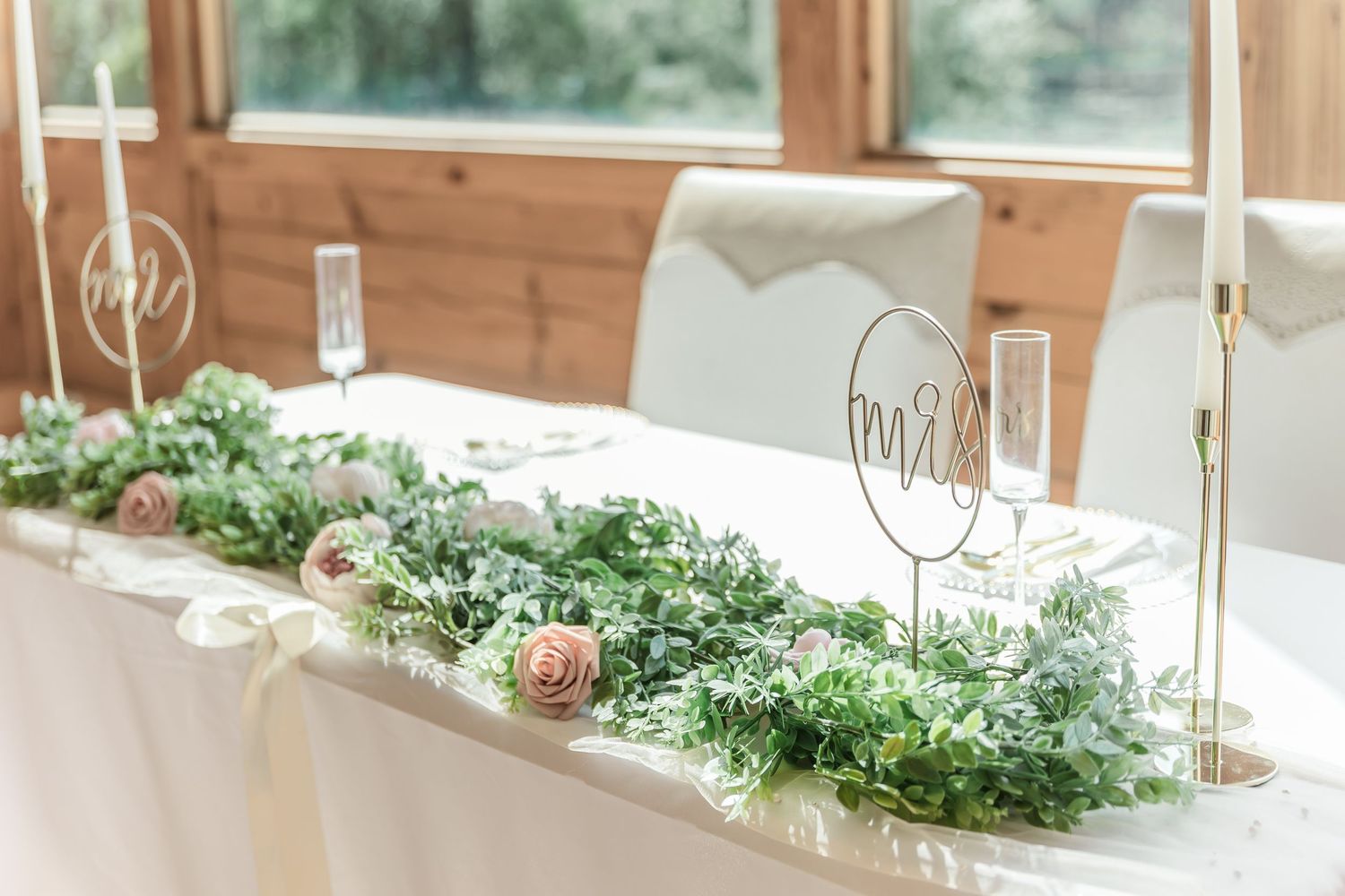 Wedding reception table with lush greenery runner, white candles, and metallic monogram signs.