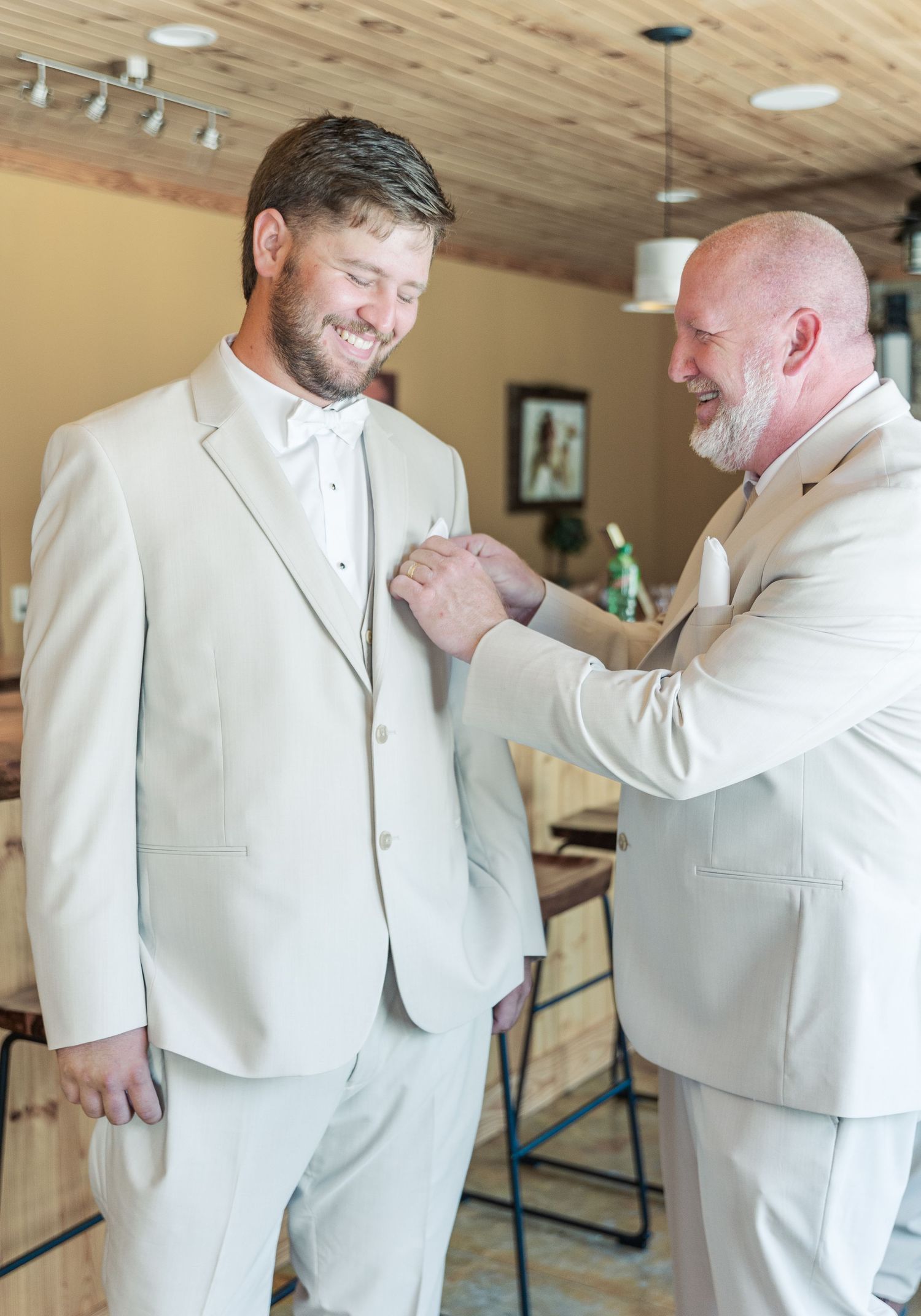 Two men in white suits share a moment while adjusting formal attire.